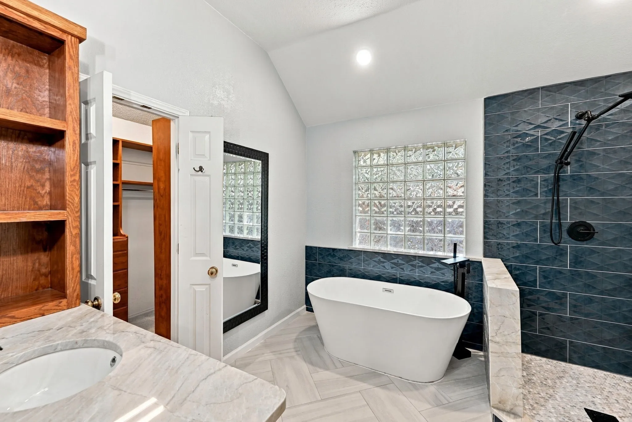 Freestanding soaking tub, walk-in shower with textured tile surround, glass block window, wood-finish shelving, and vanity with integrated sink
