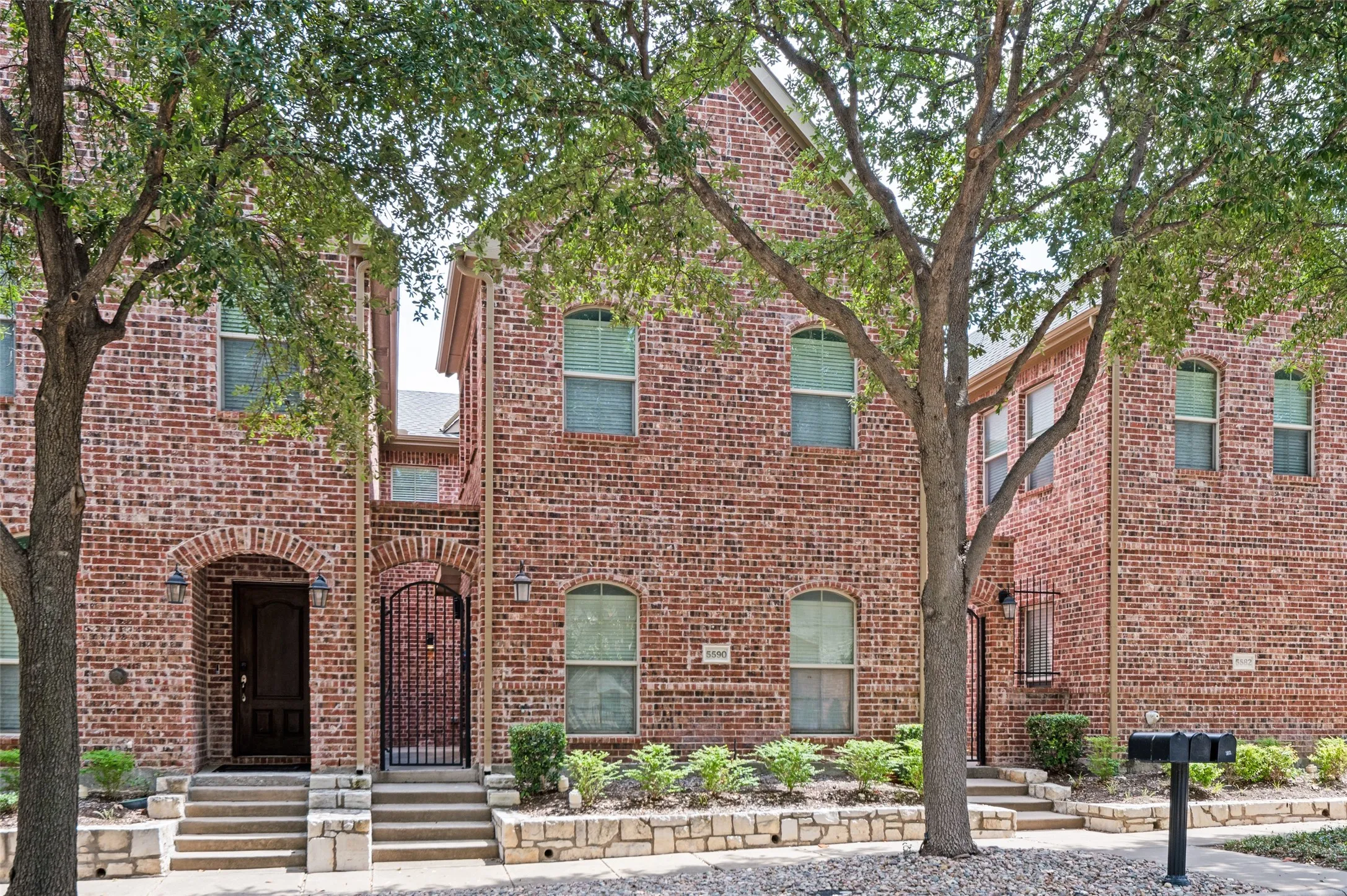 Brick exterior with arched entryways, stone-edged landscaping, mature trees, and a paved walkway