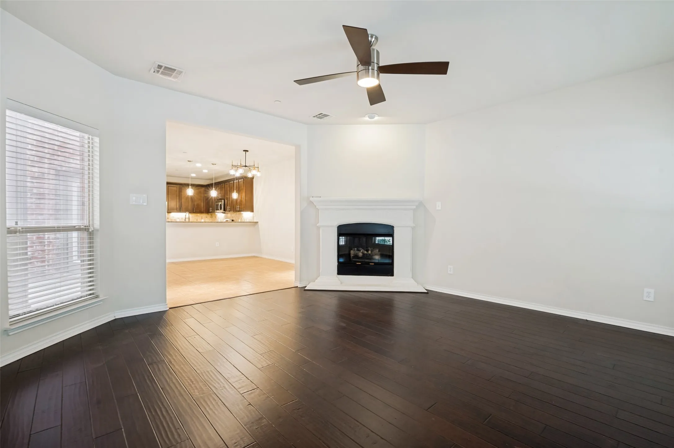 Dark wood-finish flooring, white fireplace with mantle, ceiling fan with integrated lighting, and a bay window looking into Dining Room and kitchen.