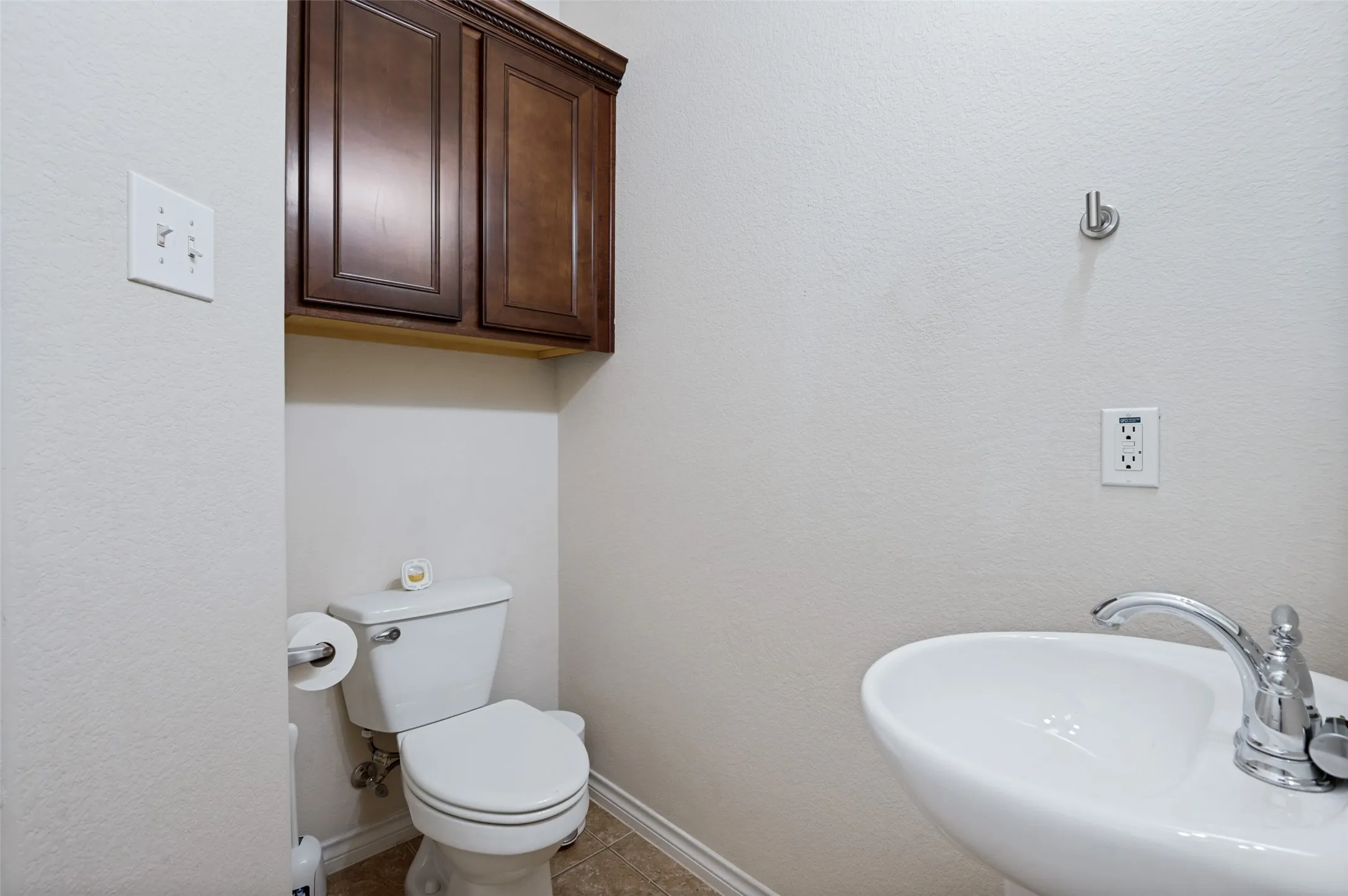 Downstairs powder bath with pedestal sink with chrome faucet, toilet, wall-mounted cabinetry, and tile flooring