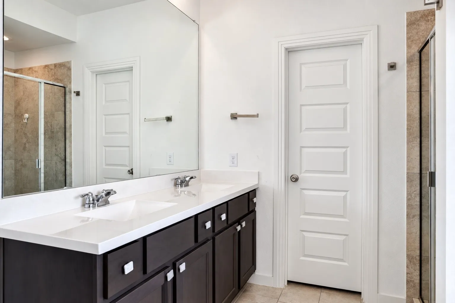 Dual sinks with chrome fixtures, expansive vanity with white surface, dark wood-finish cabinetry with silver-tone hardware, full-width mirror, and a glass-enclosed shower