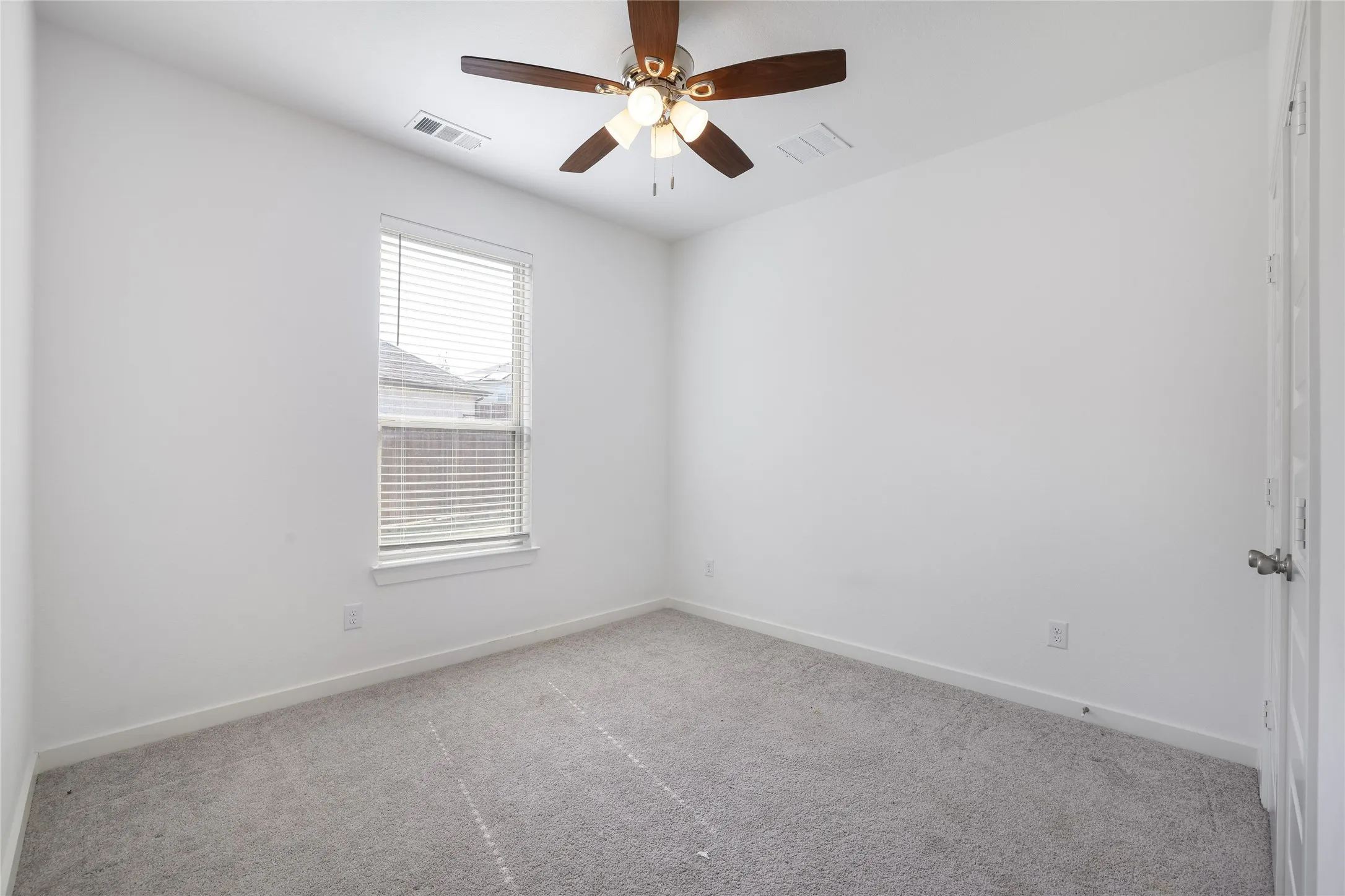 Neutral-toned carpeting, ample natural light from a window with horizontal blinds, and a ceiling-mounted fan with integrated lighting
