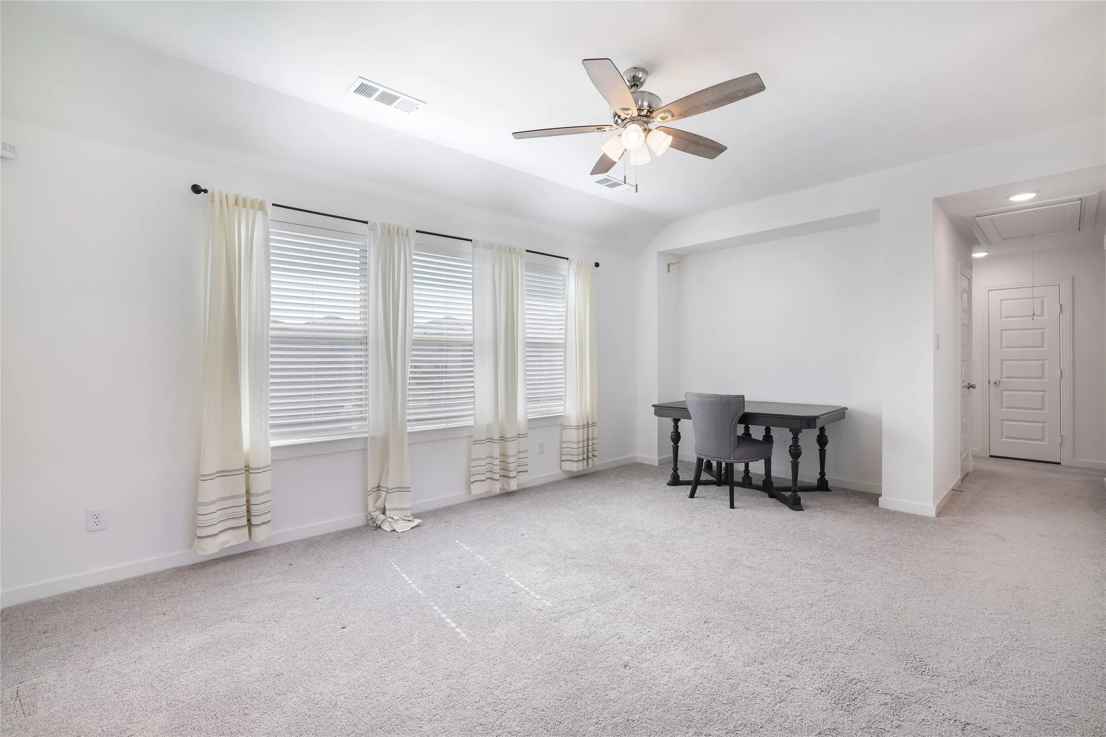 Expansive room with neutral carpeting, three windows with blinds, a ceiling fan with integrated lighting, and white walls