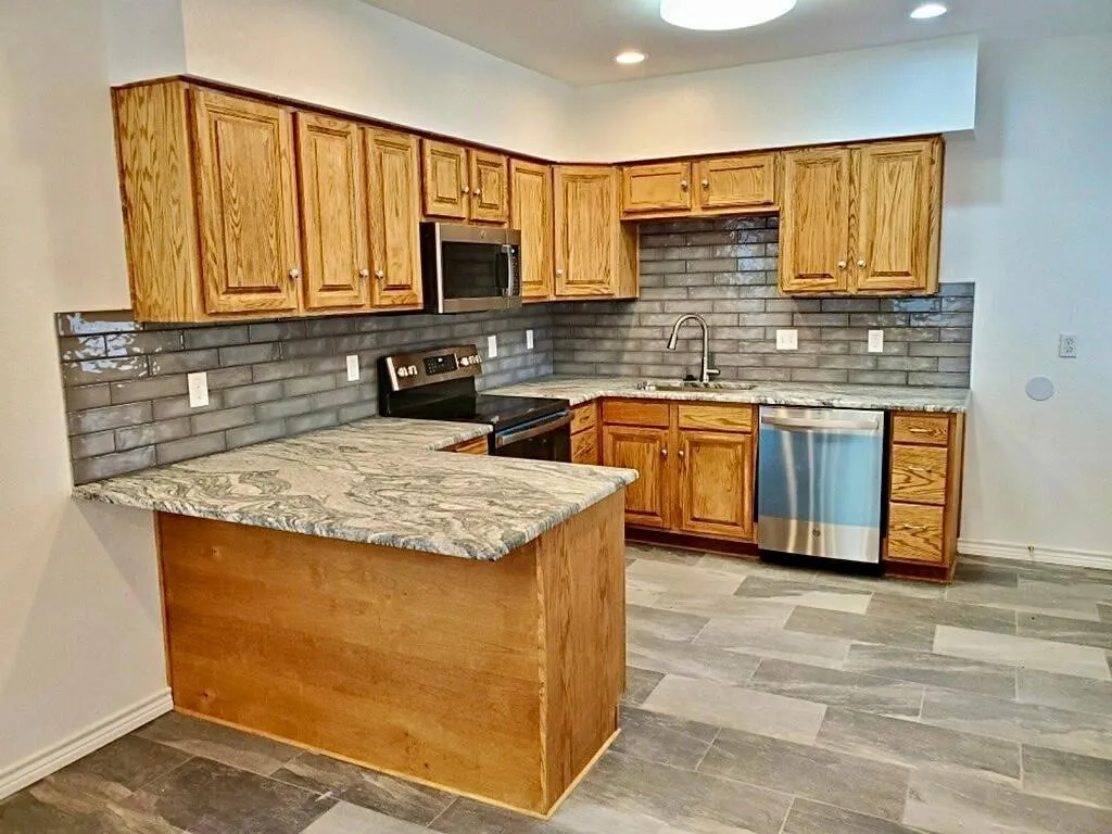 U-shaped kitchen featuring wood cabinetry, integrated stainless steel appliances, grey subway tile backsplash, and light-toned countertops