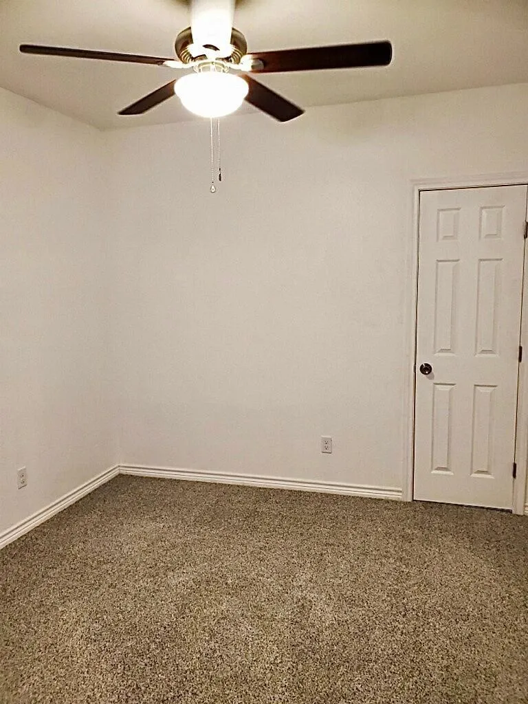 Neutral tone wall paint, textured carpet flooring, ceiling fan with integrated lighting, and a paneled interior door