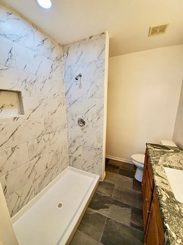 Walk-in shower with tiled surround and built-in niche, dark stone-finish floor tiles, and a vanity with a granite countertop and integrated sink