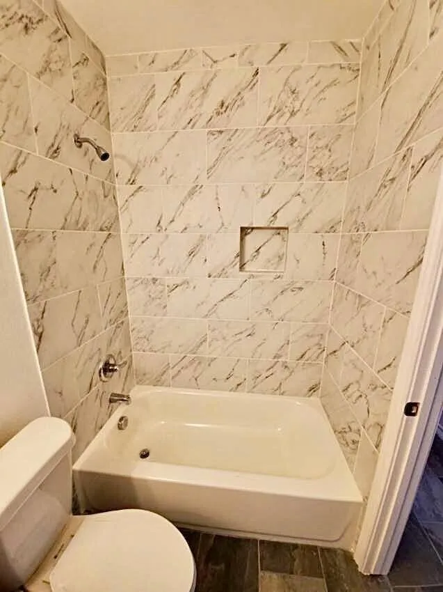 Bathtub with chrome fixtures, tiled shower surround featuring a built-in niche, and dark-toned floor tiling