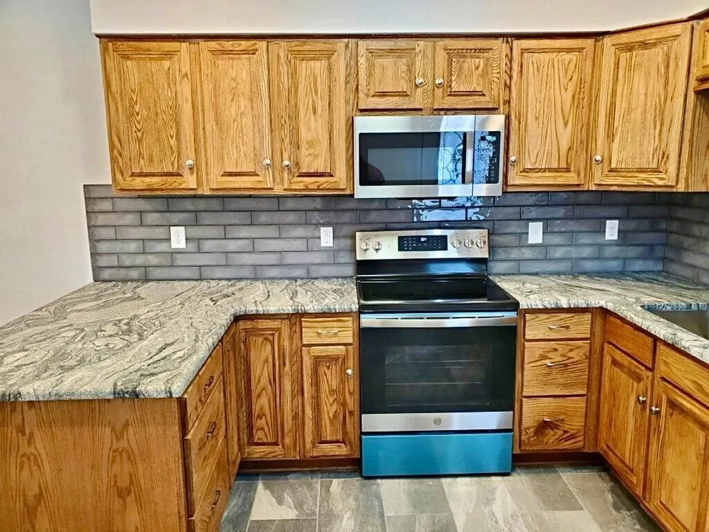 Wood cabinetry, patterned countertops, brick-patterned backsplash, stainless steel microwave, and a stainless steel oven/range