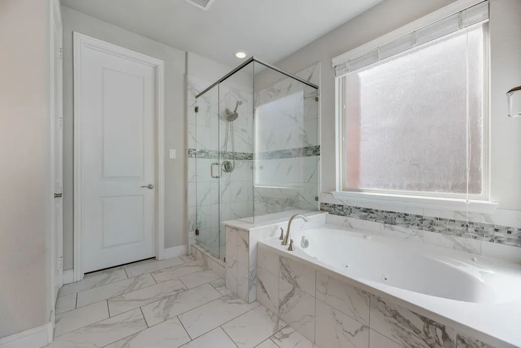 Spacious bathroom featuring a built-in soaking tub, glass-enclosed shower, white tile flooring, and a decorative tile accent band