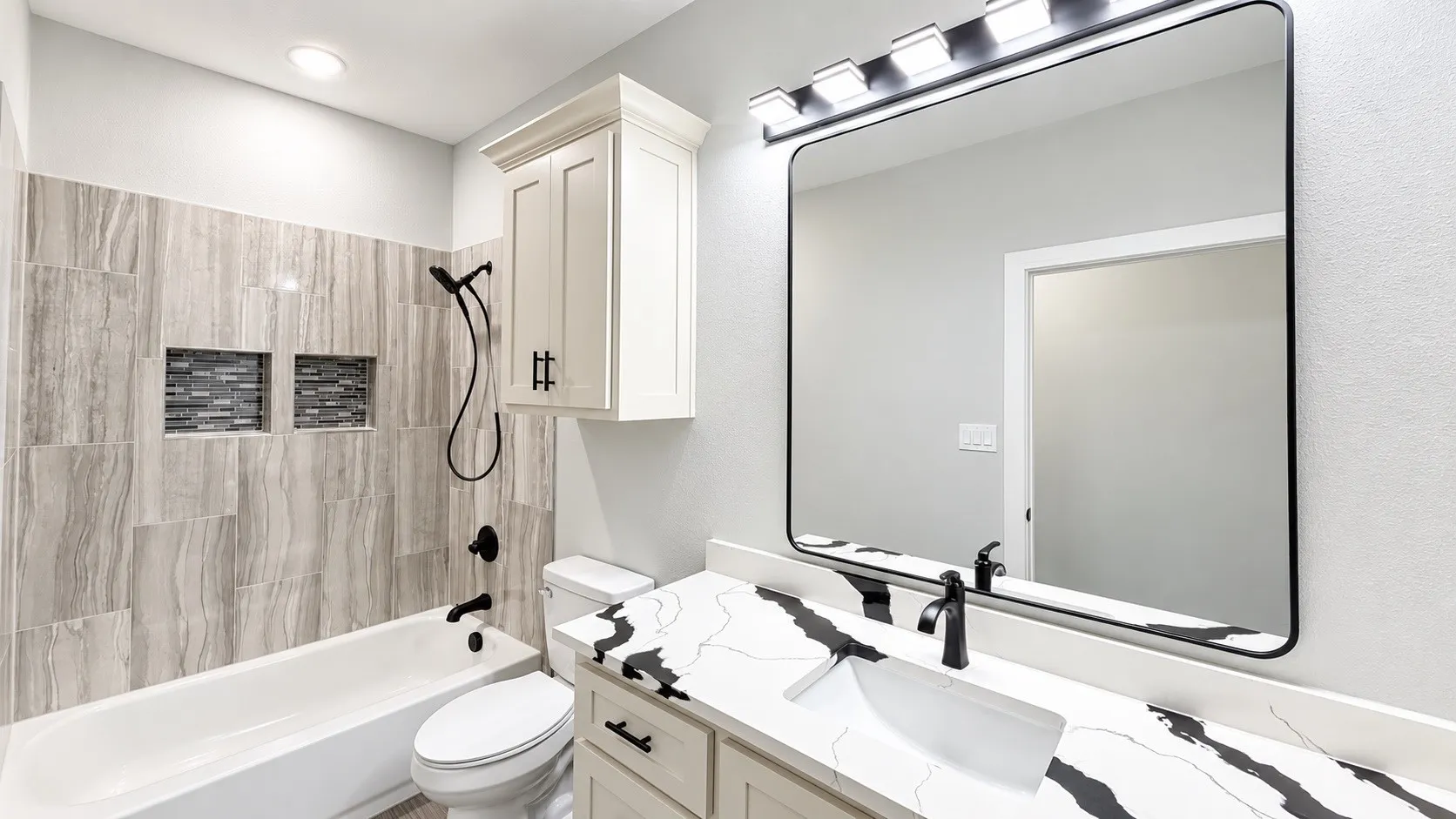 Modern bathroom featuring a soaking tub with tiled surround, dual shower niches with mosaic tile, a vanity with a patterned countertop, a rectangular mirror, and a wall-mounted light fixture