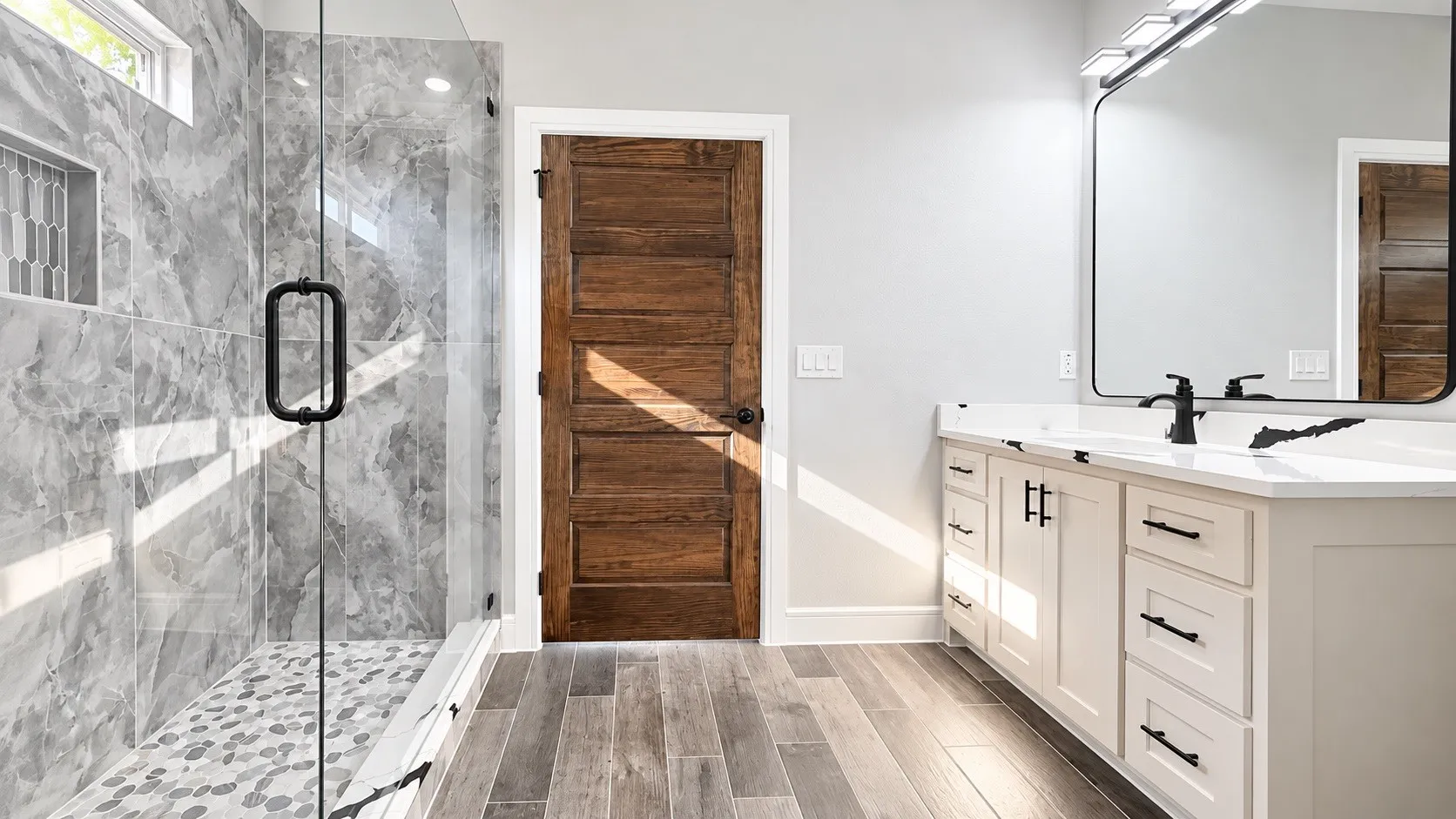 Glass-enclosed shower with gray marbled tile surround, pebble shower floor, white vanity with black hardware, white countertop, and wood-finish flooring