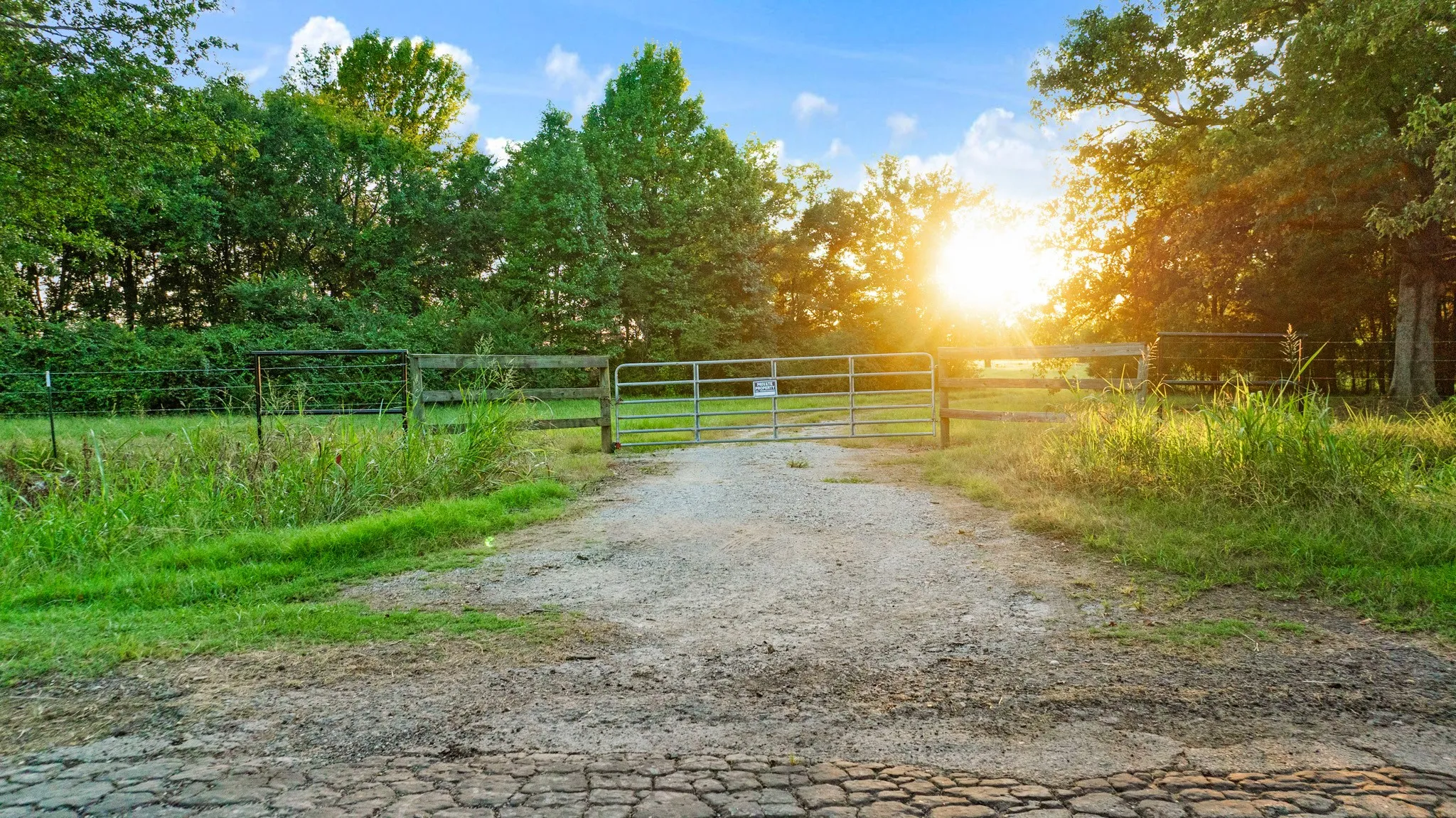 Gated gravel driveway leading to a treed property with barbed wire fence boundary
