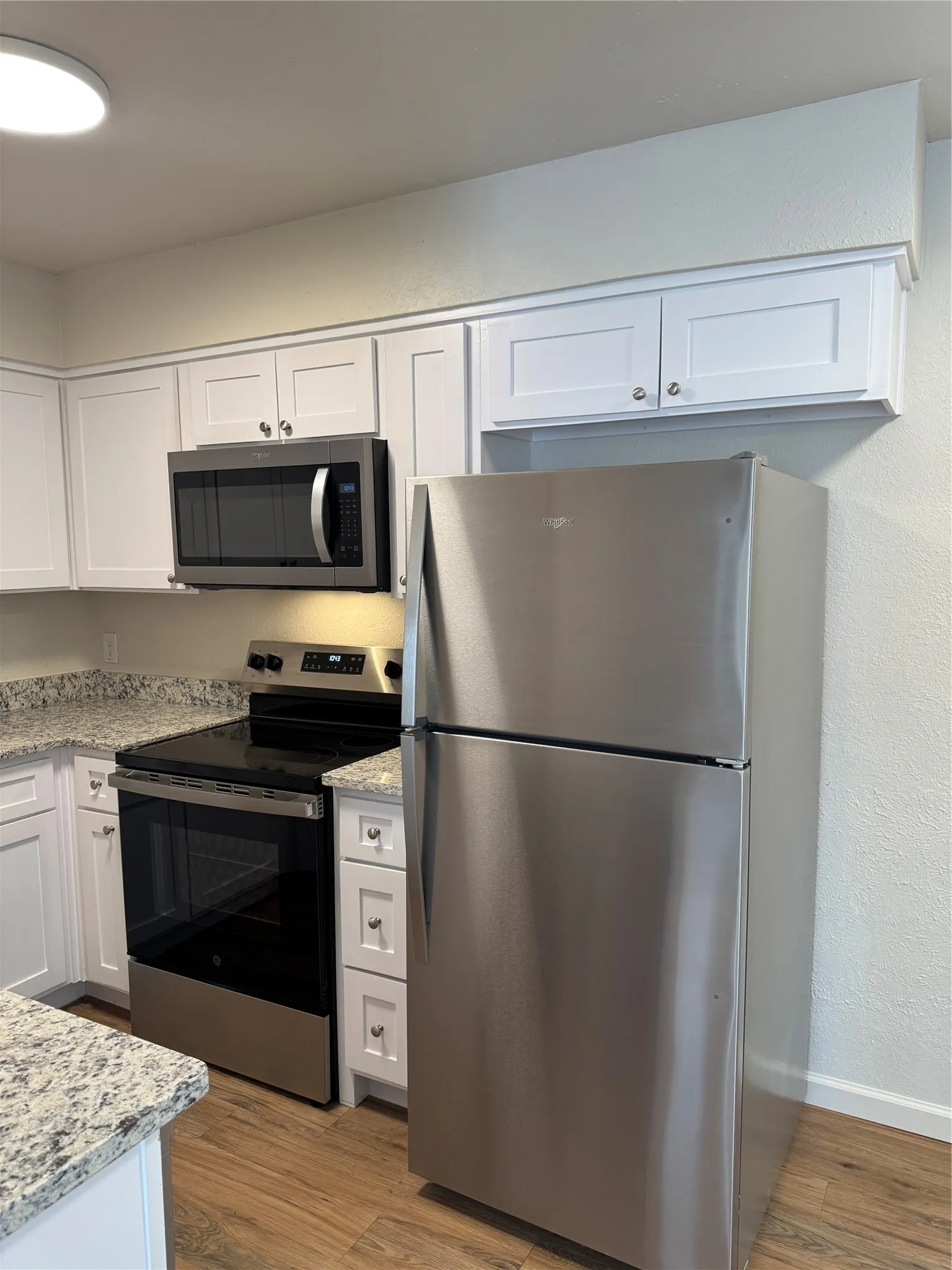 Stainless steel appliance suite, white shaker cabinetry, speckled countertops, wood-finish flooring, and recessed lighting