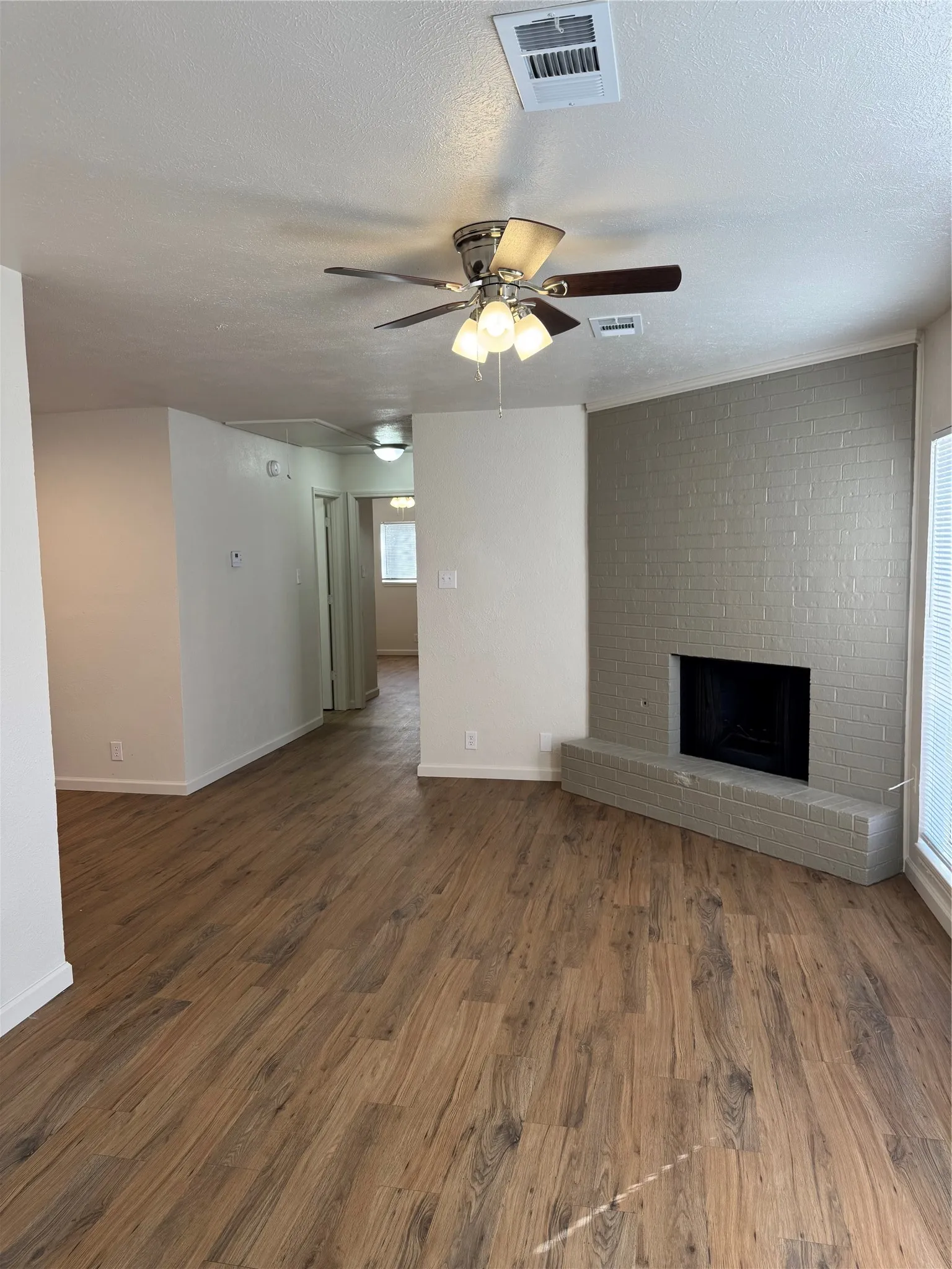 Wood-finish flooring throughout, brick-surround fireplace with hearth, ceiling fan with integrated lighting, and white walls