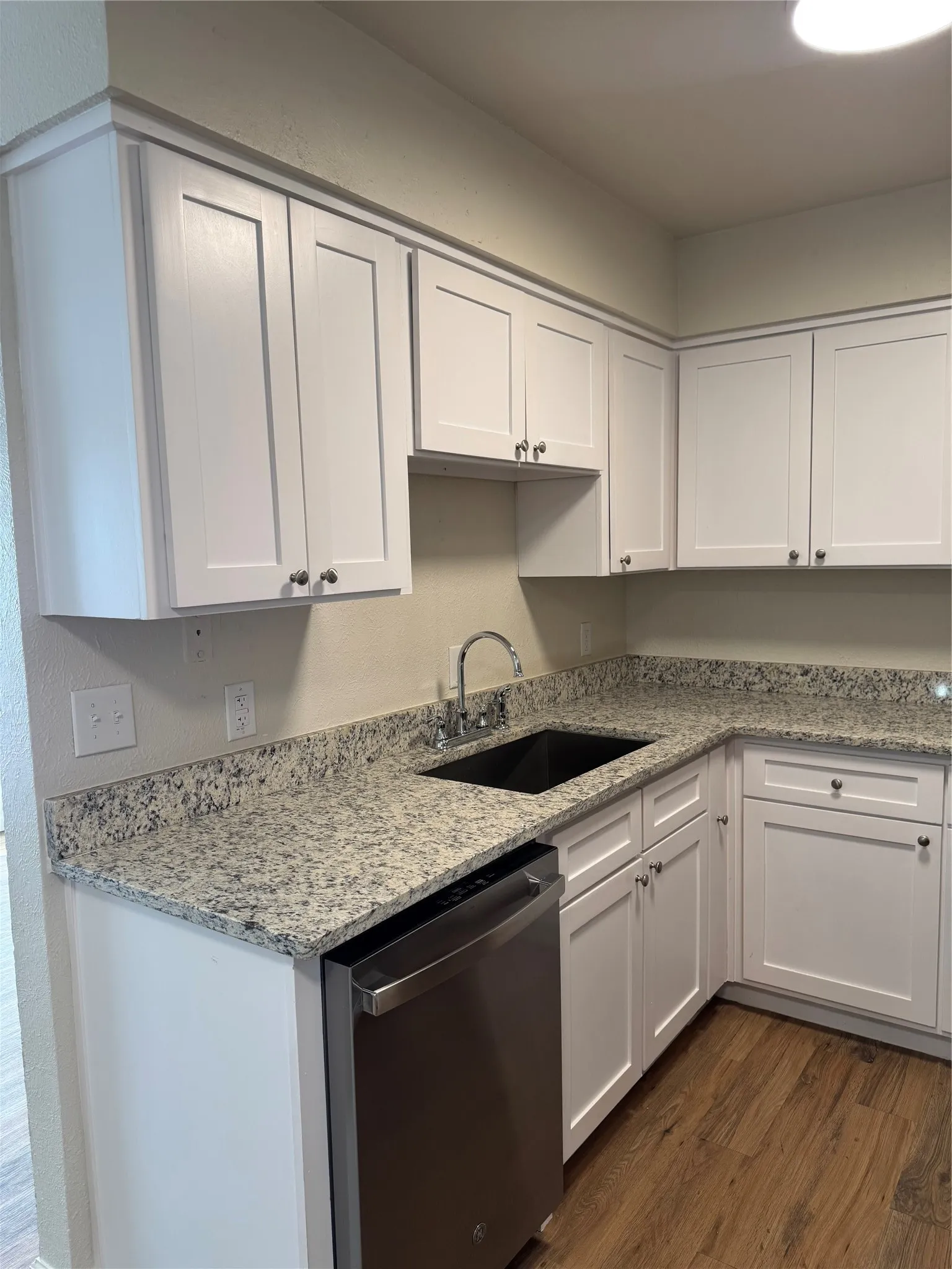 White shaker-style cabinetry, patterned stone countertops, stainless steel dishwasher, undermount sink with gooseneck faucet, and wood-finish flooring