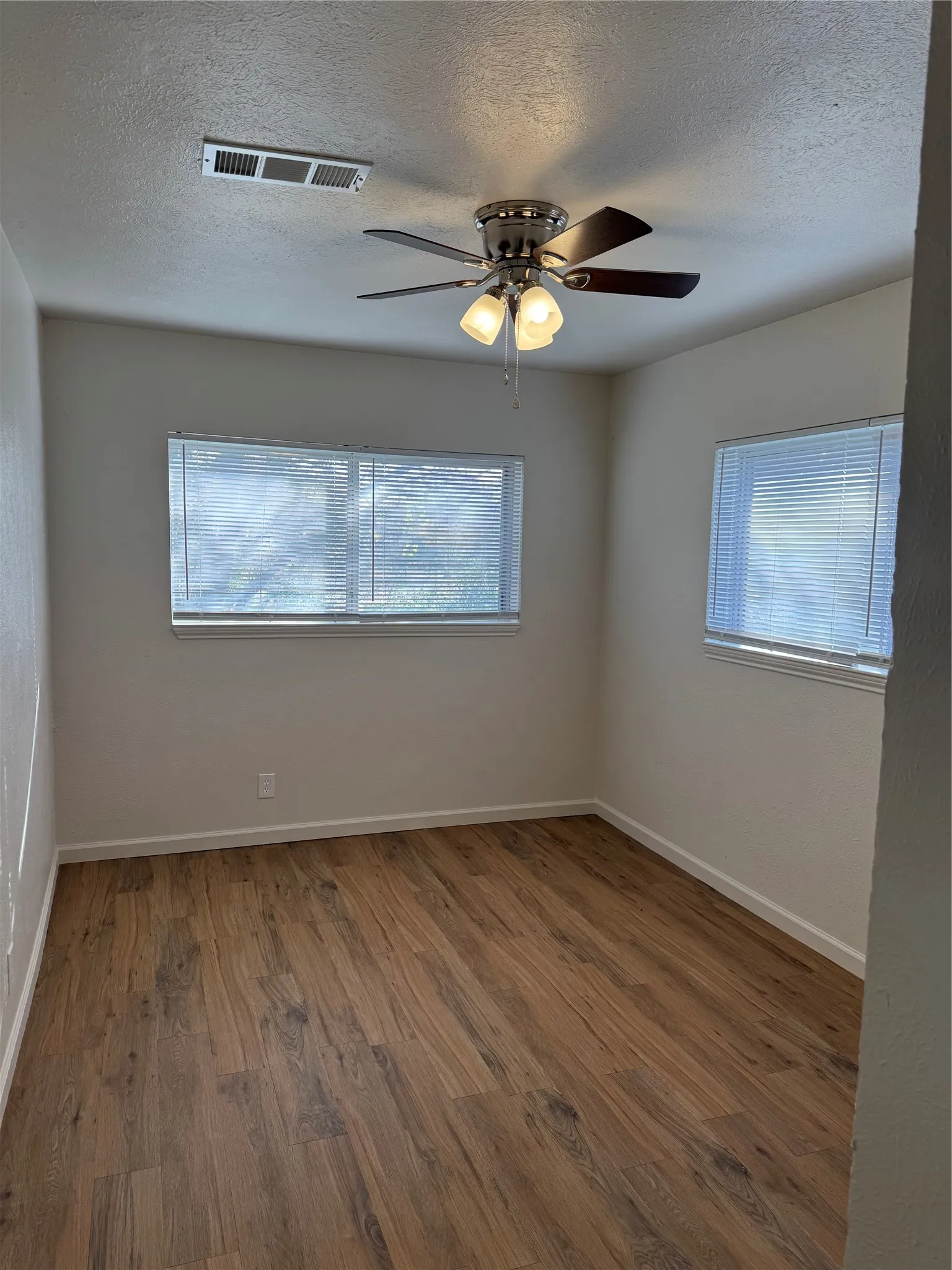 Wood-finish flooring, white walls, two windows with blinds, and a ceiling fan with integrated lighting