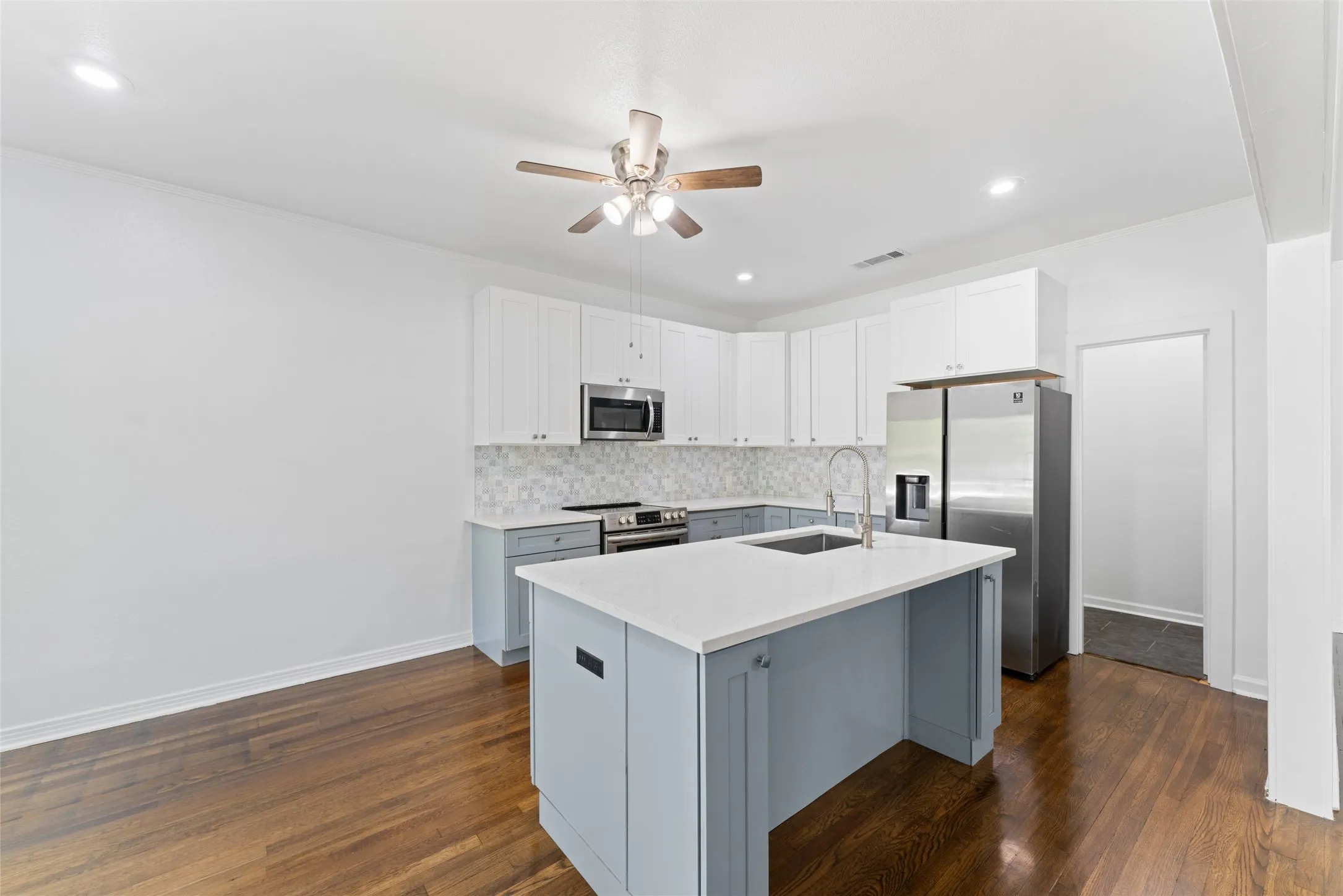 Modern kitchen featuring a central island with a sink, stainless steel appliances, upper white cabinetry, lower blue cabinetry, and wood-finish flooring