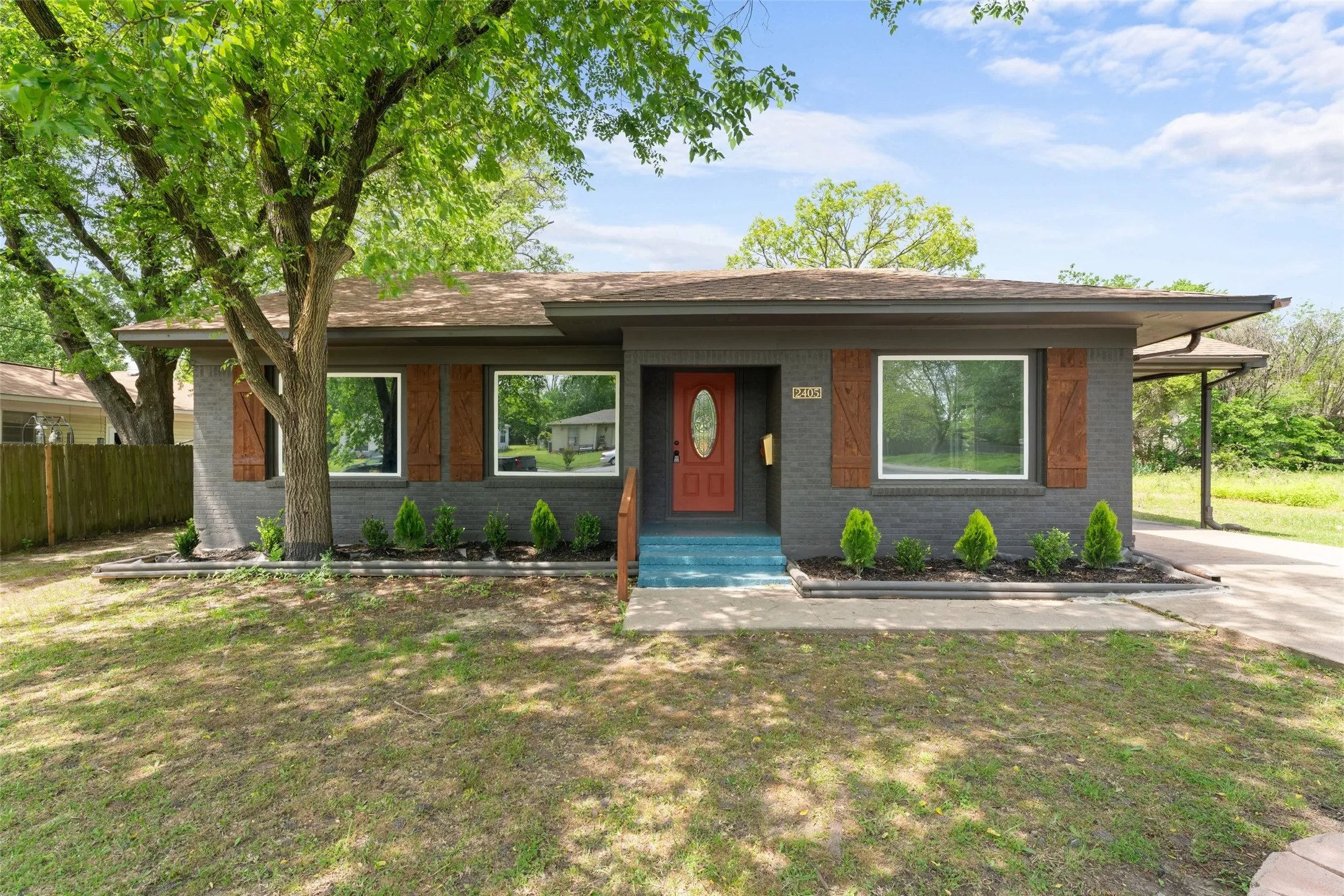 Exterior featuring a dark gray brick facade, a vibrant red entry door, a carport, and a landscaped front planting bed