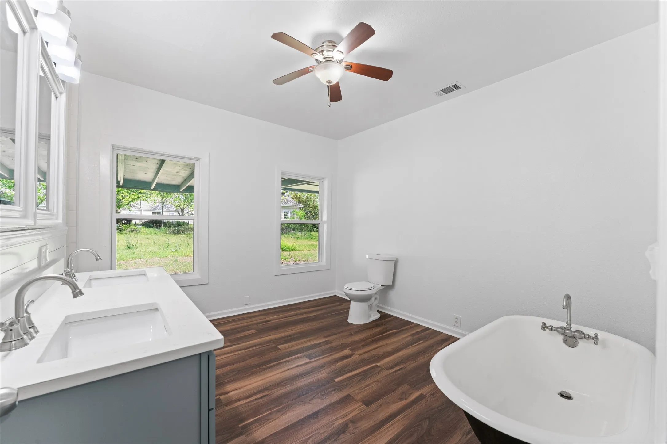 Dual vanity with undermount sinks, wood-finish flooring, freestanding bathtub, and ceiling fan with lighting