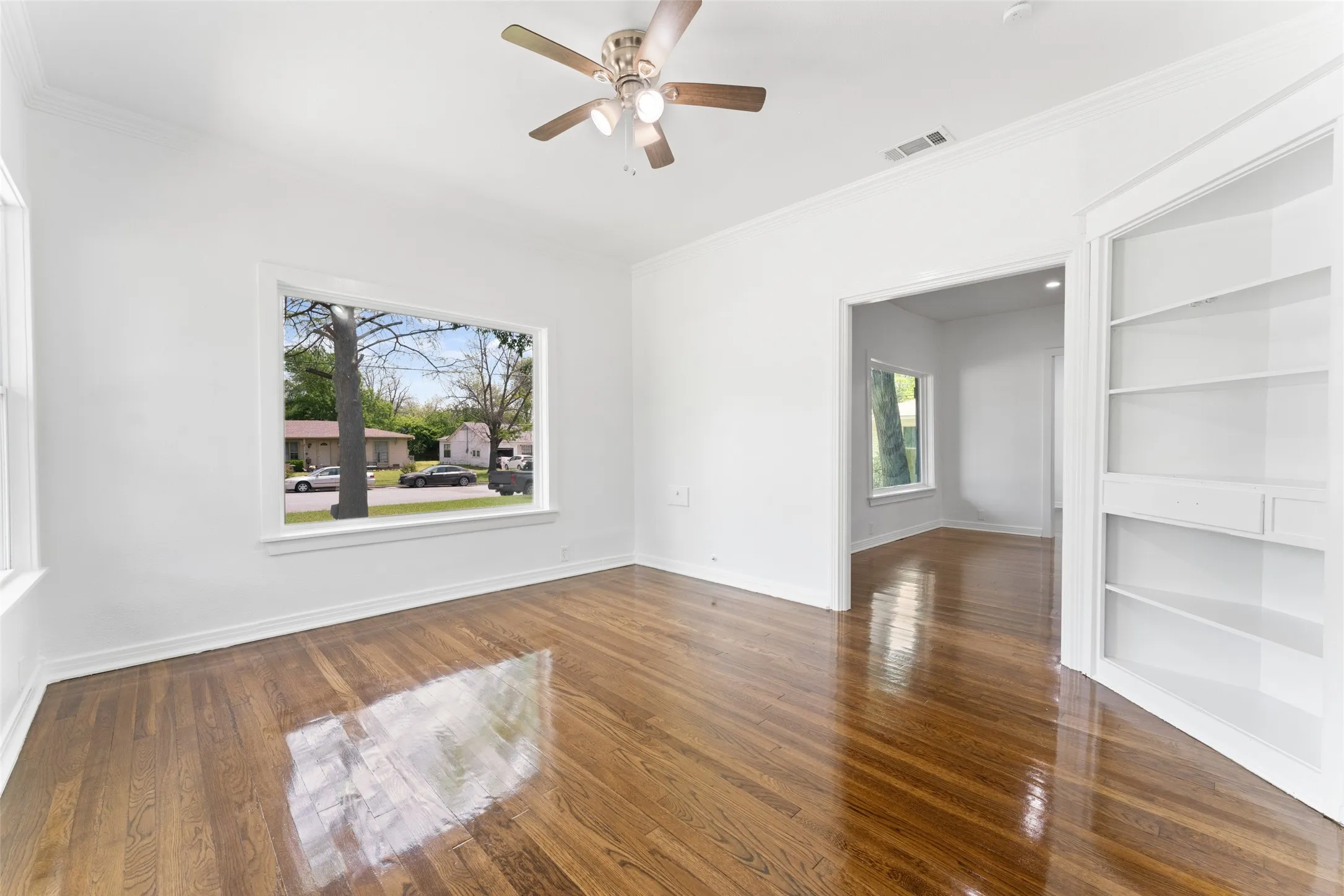 Polished wood flooring, white painted walls, ceiling fan with light fixture, large window, and built-in shelving