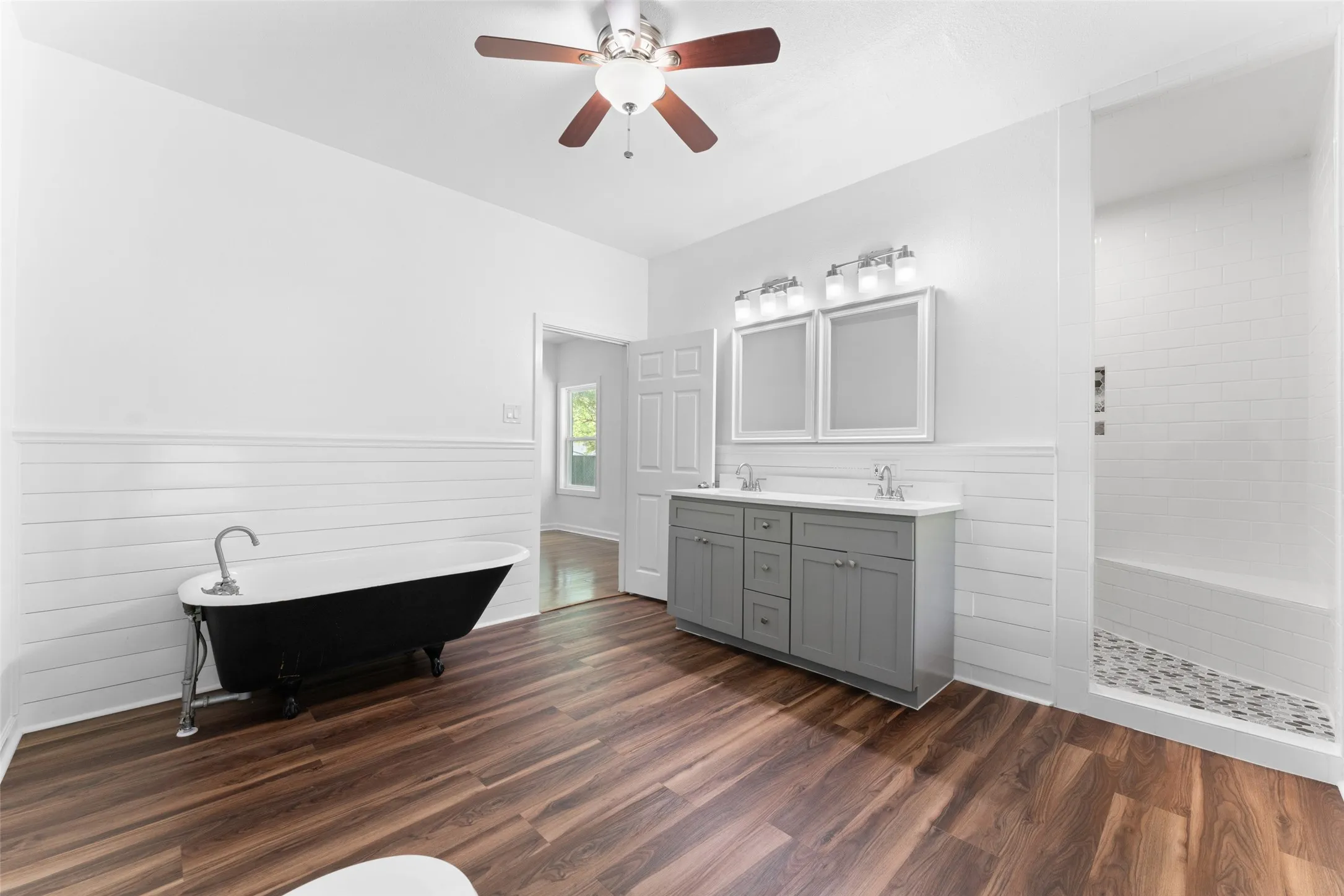 Wood-finish flooring, freestanding clawfoot tub, dual vanity with shaker-style cabinetry, walk-in shower with subway tile, and a ceiling fan