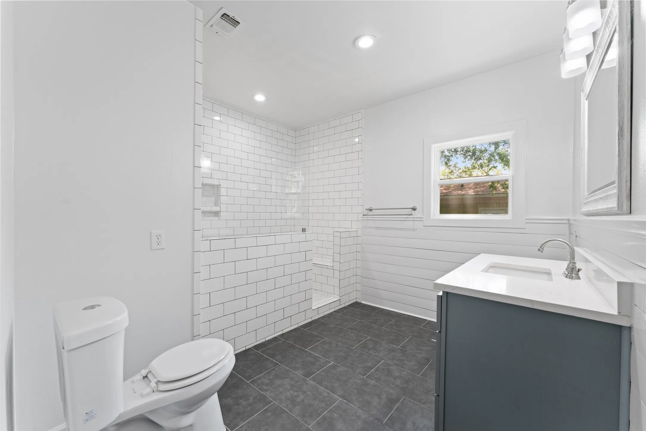 White subway tile shower with glass enclosure, single vanity with integrated sink, dark gray tile flooring, and white shiplap wainscoting