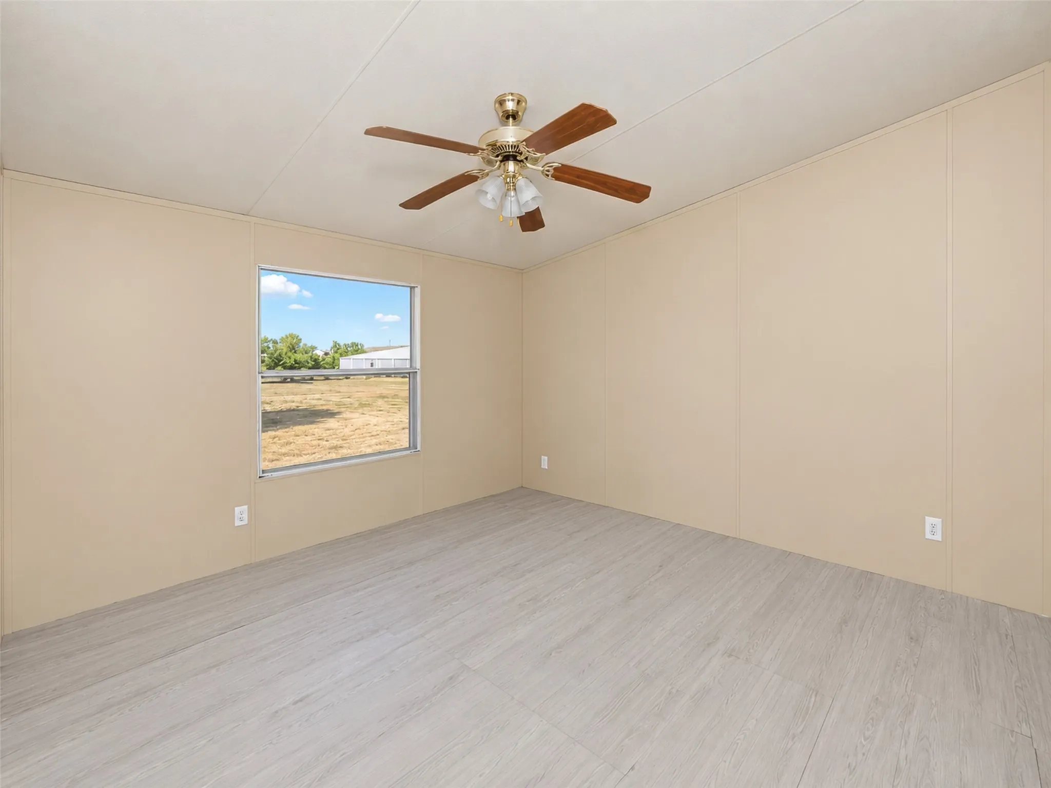 Wood-finish ceiling fan with integrated lighting, window with exterior landscape, light wood-finish flooring, and neutral wall paneling