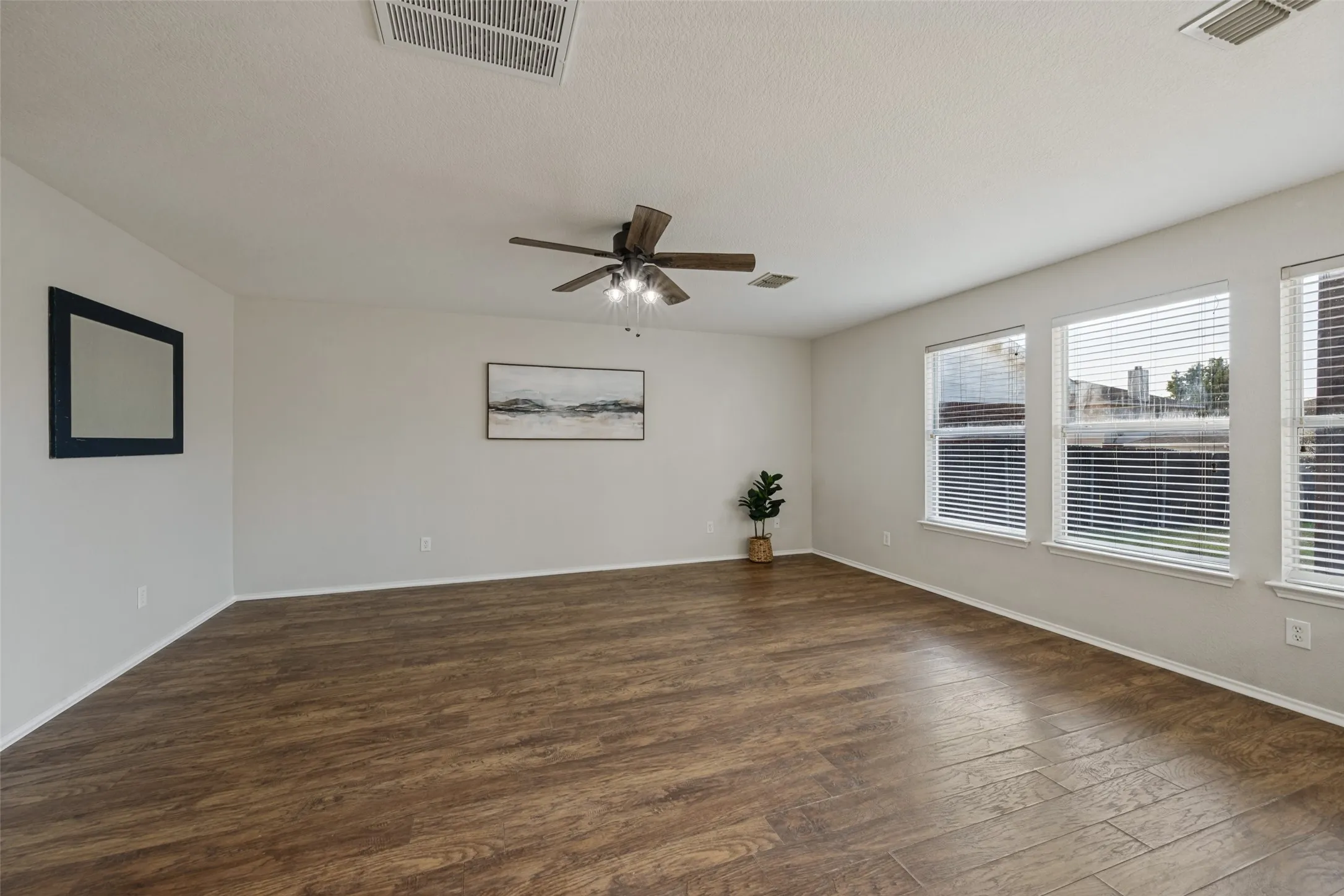 Expansive living area featuring wood-finish flooring, a ceiling fan with integrated lighting, multiple windows with blinds, and light-toned wall finishes