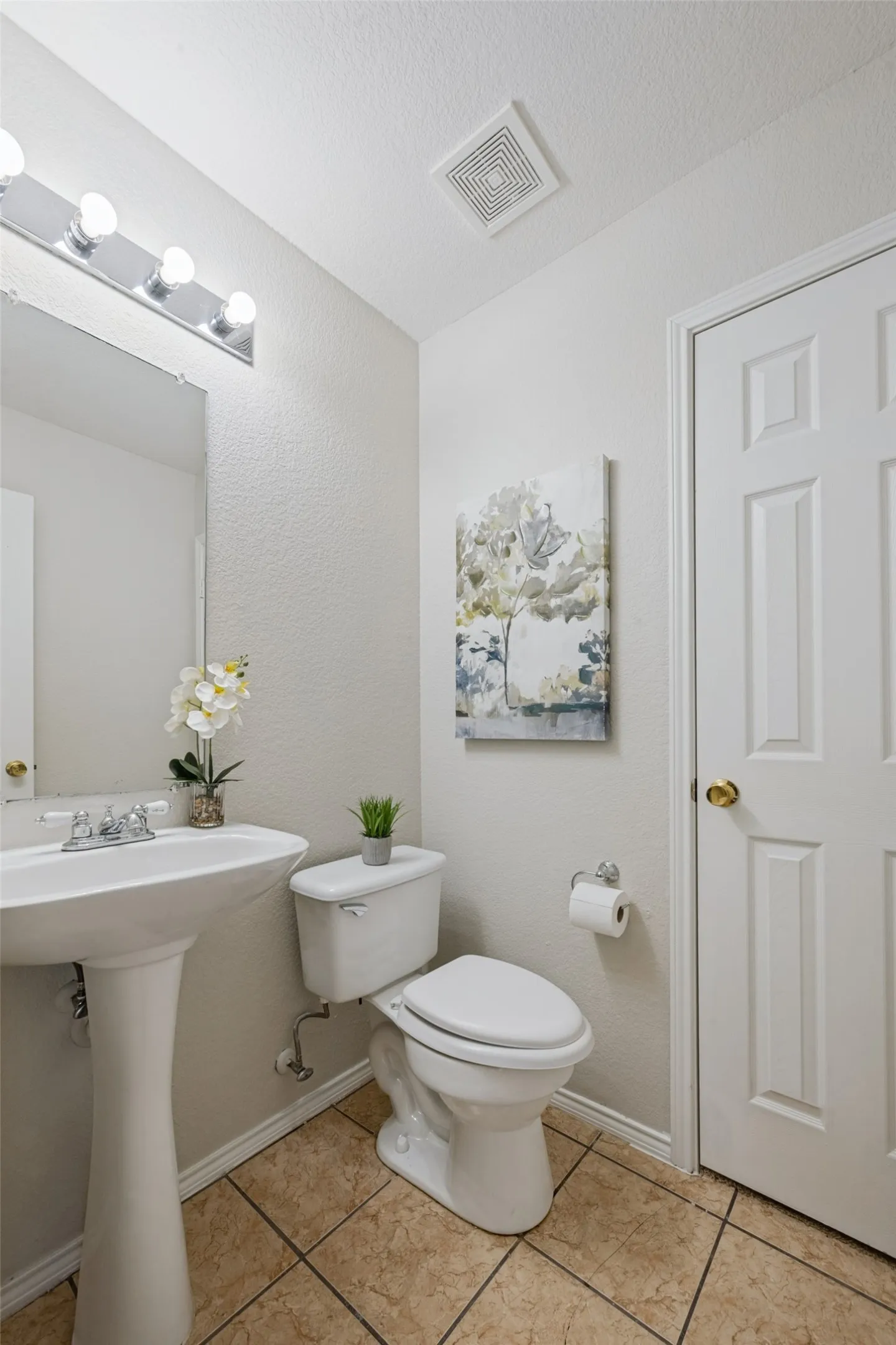 Pedestal sink with chrome-finish faucet, vanity lighting, tile flooring, and paneled door with gold-tone hardware