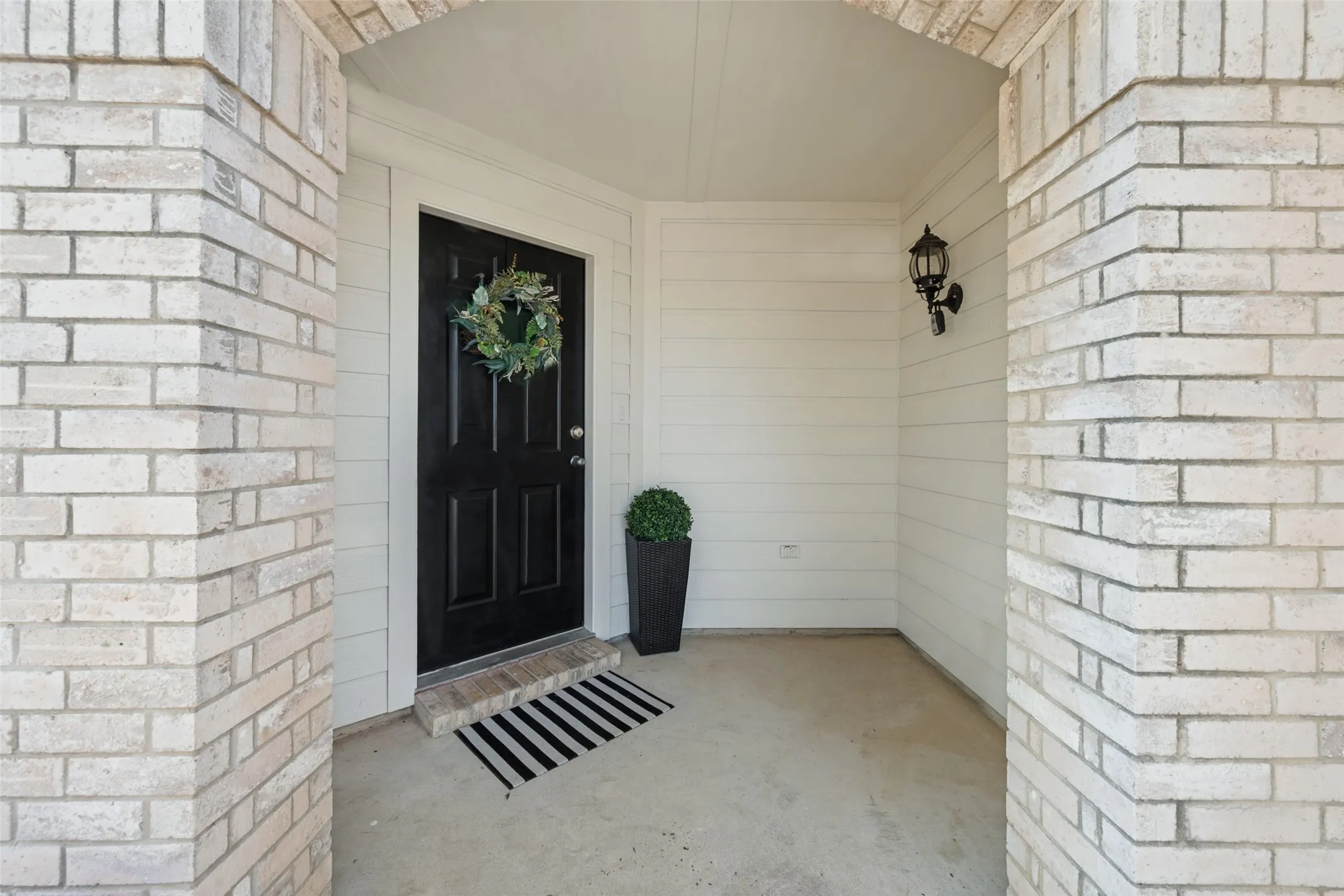 Brick columns with archway, dark panel door, transom window, and exterior wall sconce