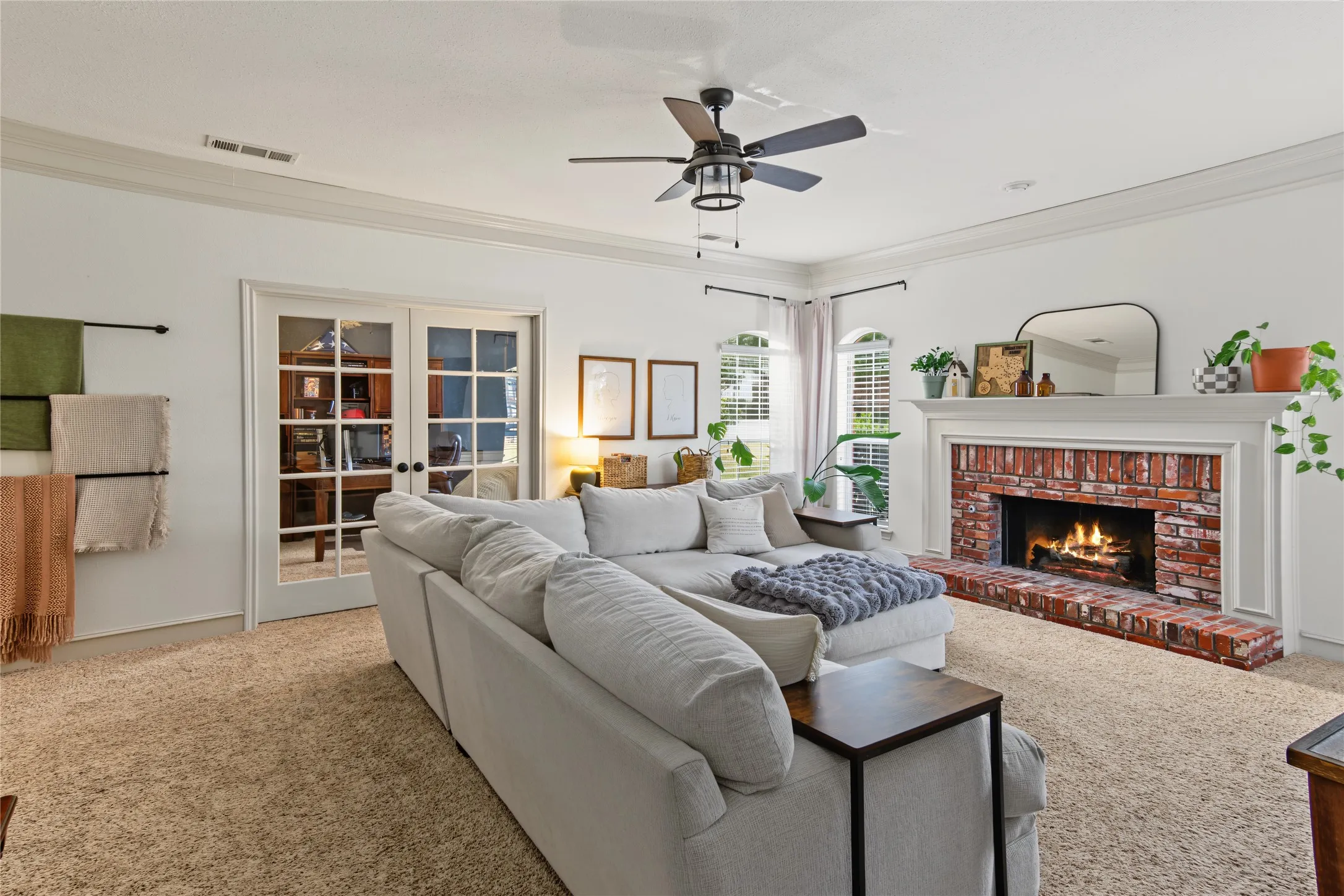 Brick-surround fireplace with a substantial white mantel, crown molding, double French doors, large arched windows, and a ceiling fan with integrated lighting