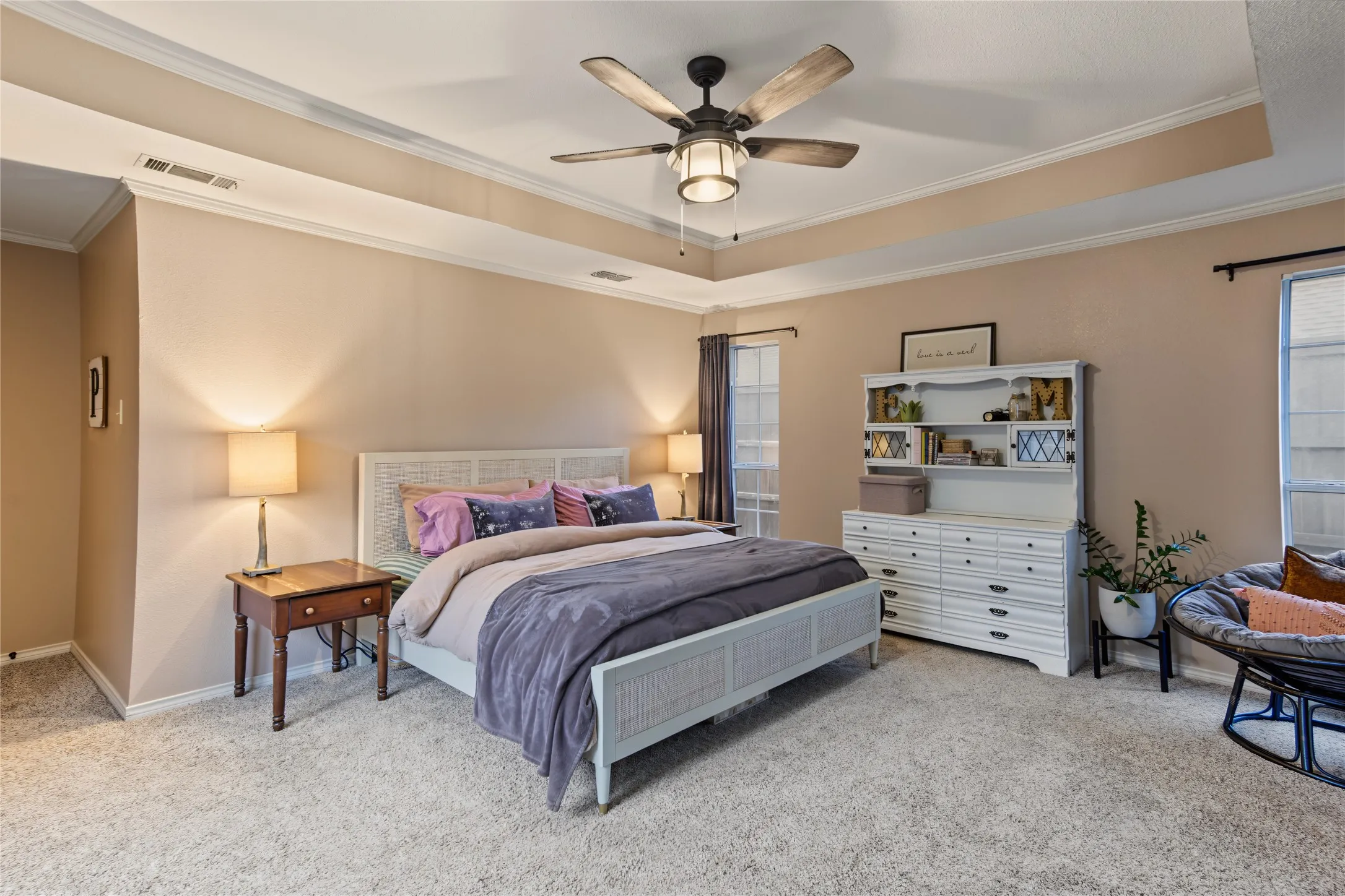 Tray ceiling with crown molding, ceiling fan with integrated lighting, two windows with blinds, and light-colored carpeting