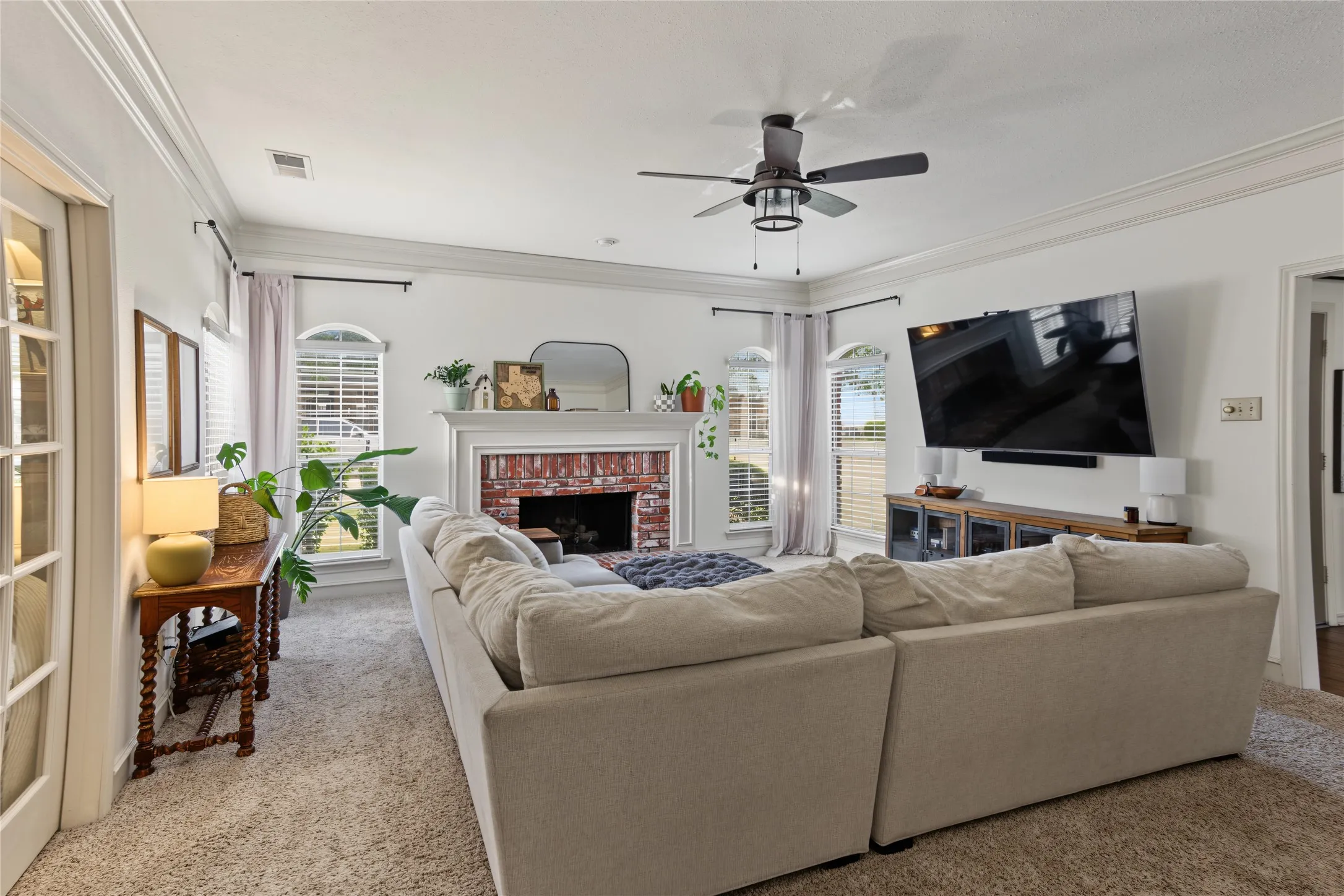 Living area featuring a brick fireplace, decorative mantel, arched windows with blinds, a ceiling fan, and crown molding
