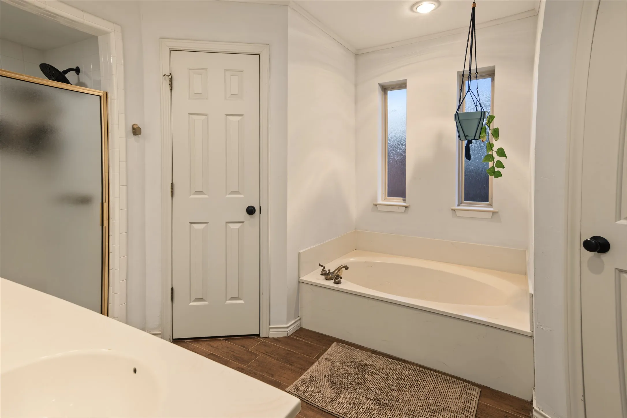 Built-in soaking tub with brass-finish fixtures, two vertical privacy windows, recessed lighting, wood-finish flooring, and a glass-enclosed shower with a black shower head