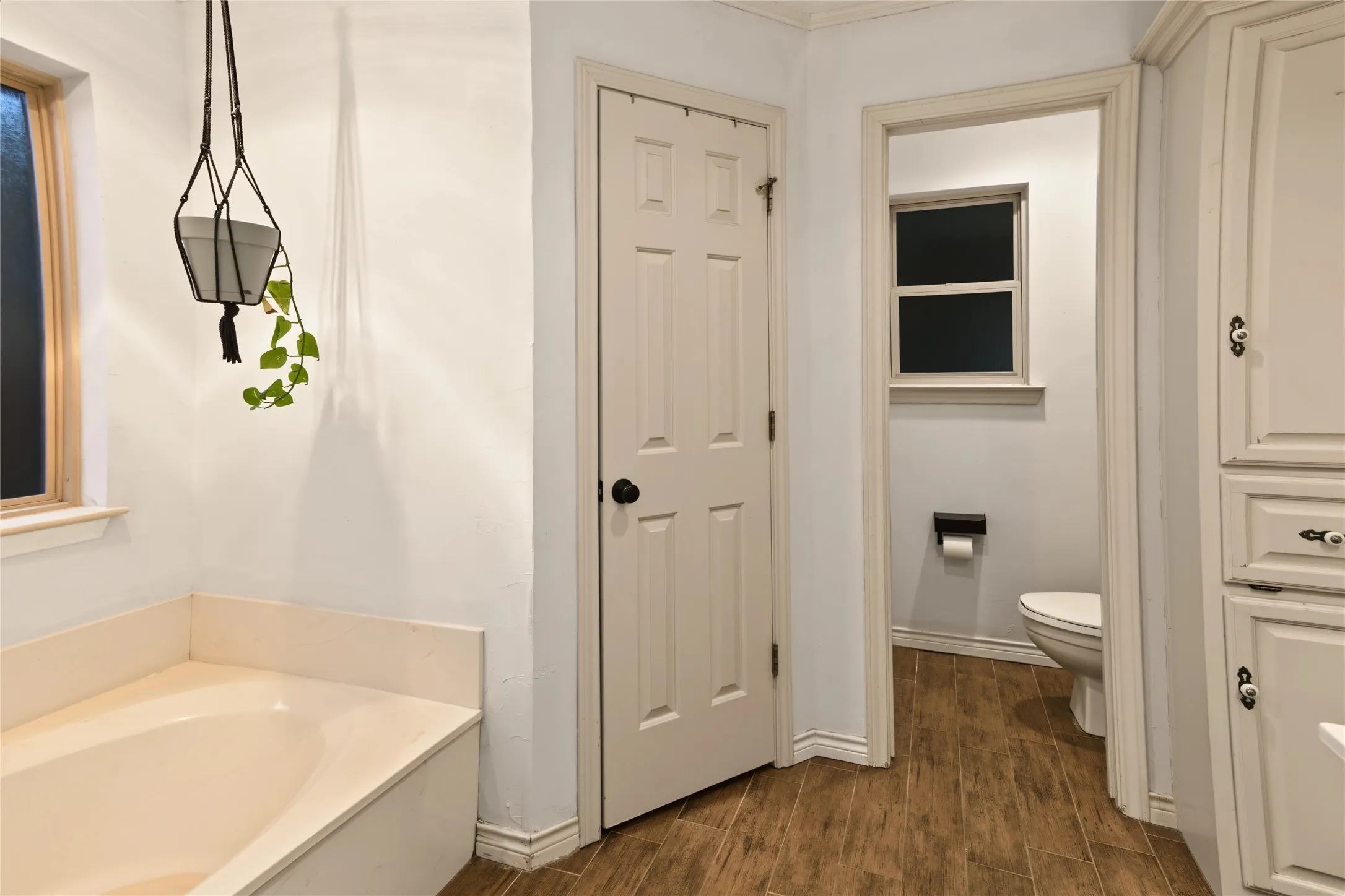 Soaking tub with a tiled surround, wood-finish flooring, and light-colored cabinetry with decorative hardware
