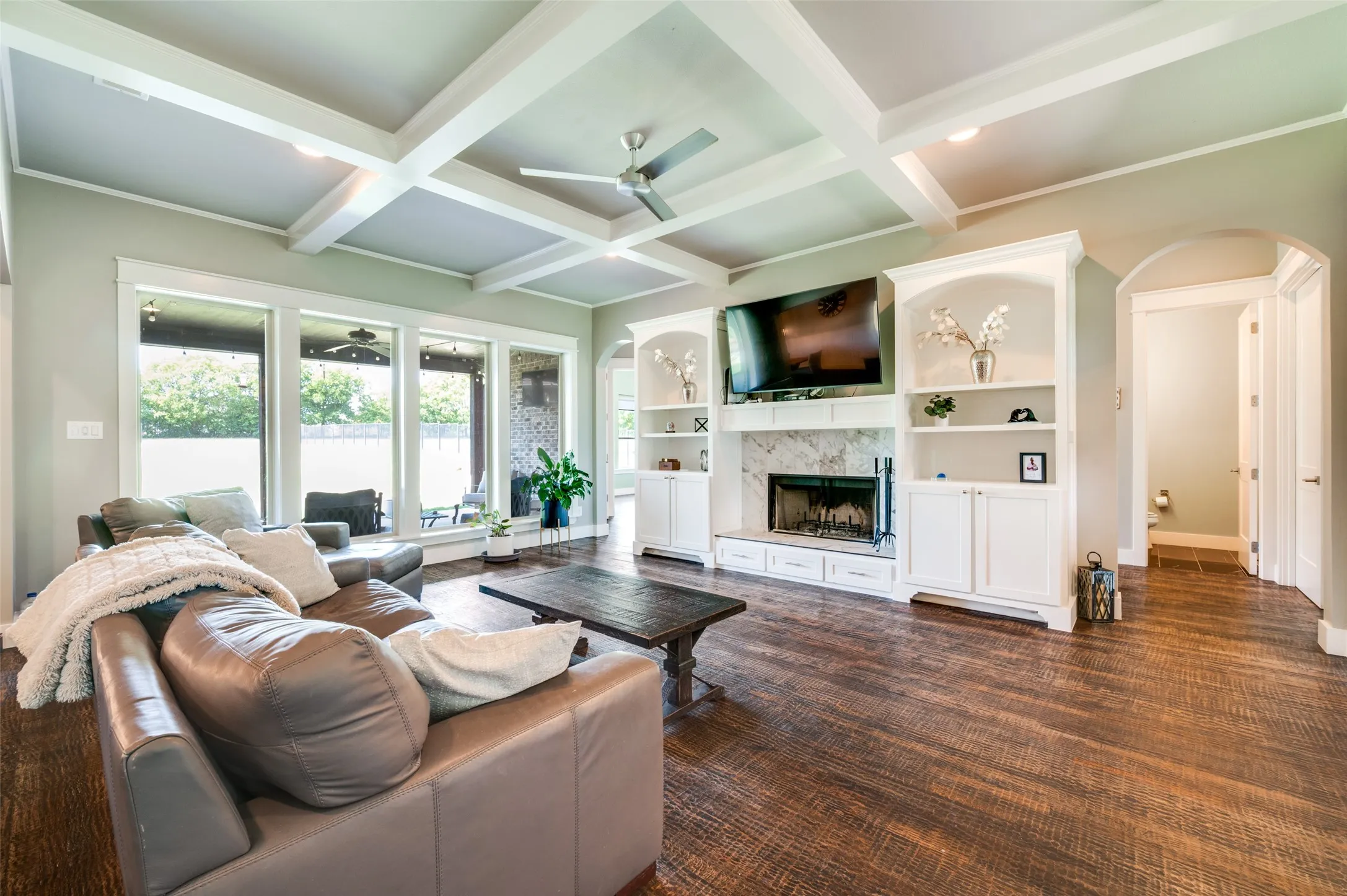Coffered ceiling details, wood-finish flooring, built-in cabinetry surrounding a fireplace, large windows, and an arched doorway