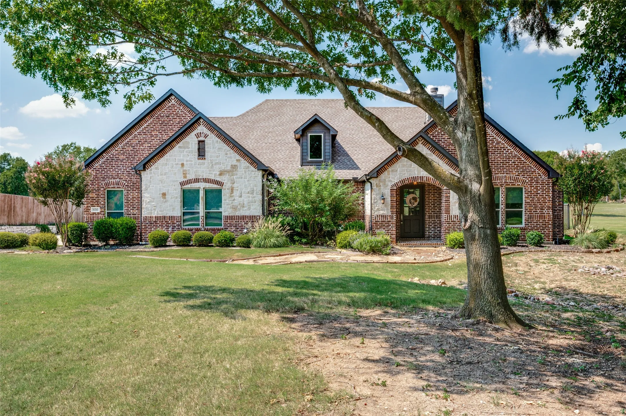 Brick and stone facade, arched entryway, gabled roofline, dormer window, and established landscaping