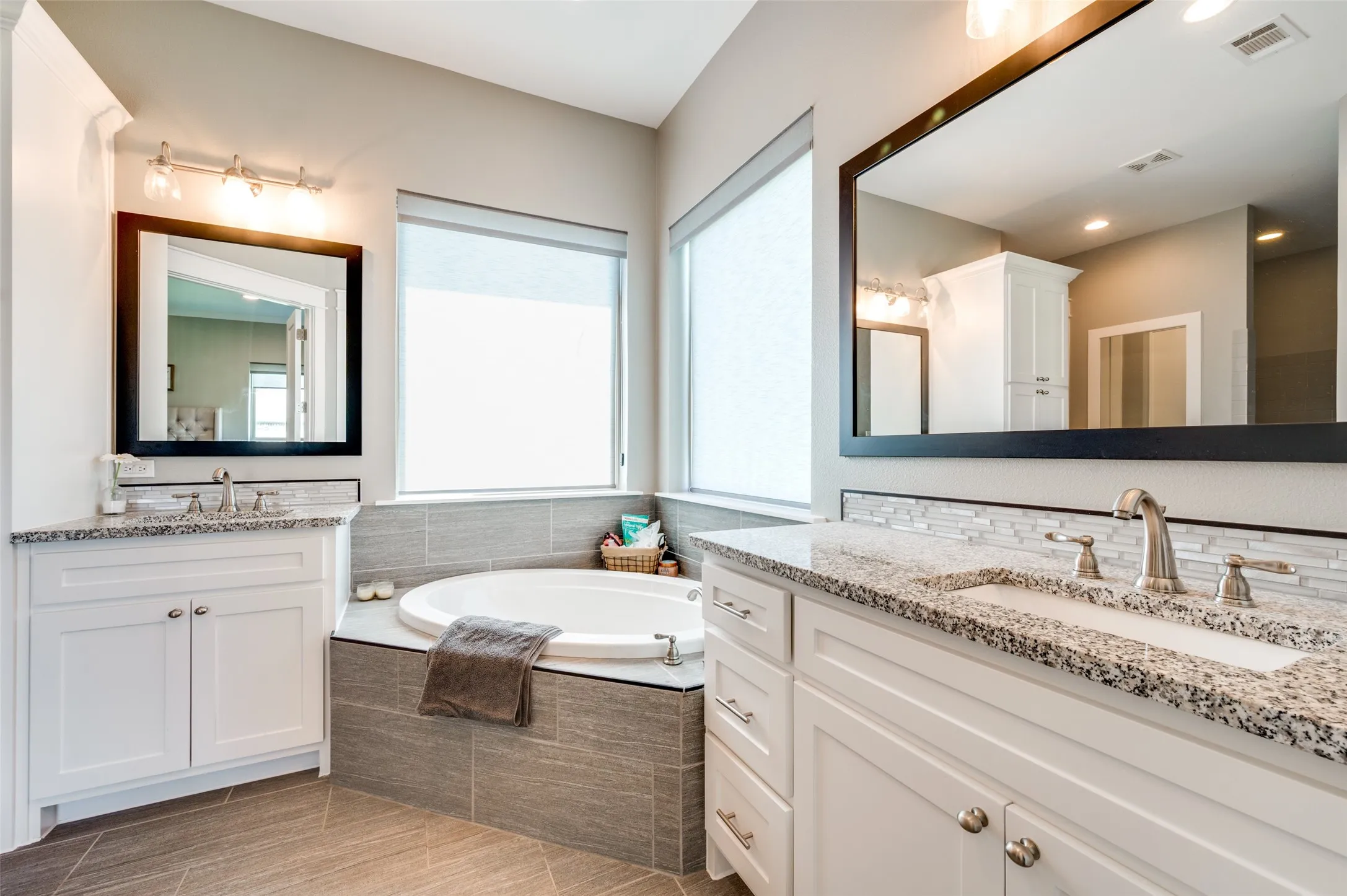 Dual white vanities with speckled countertops, a built-in soaking tub, tile flooring, and a tiled tub surround
