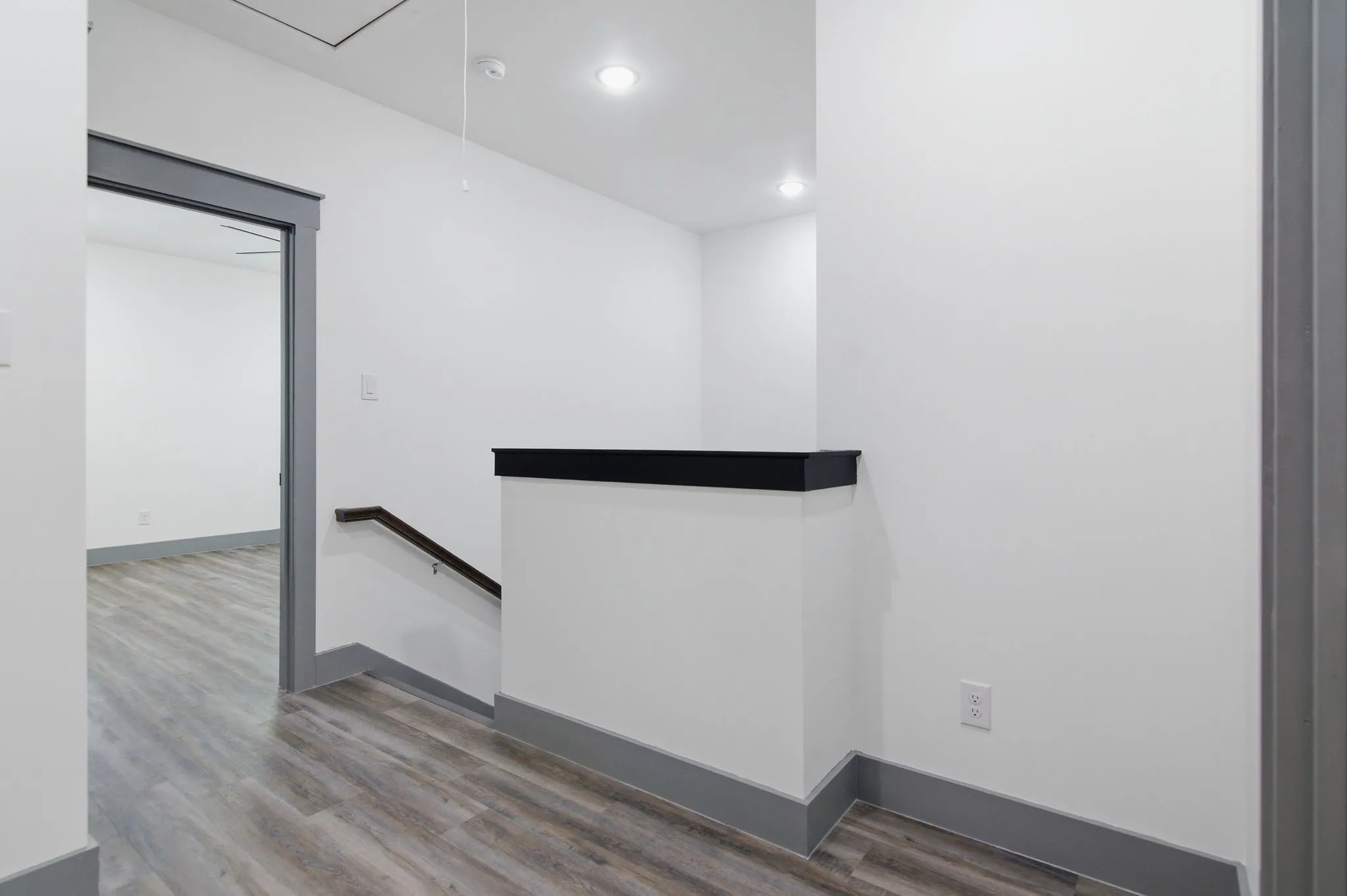 Recessed ceiling lighting, wood-finish flooring, white walls, dark gray trim, and a dark wood handrail