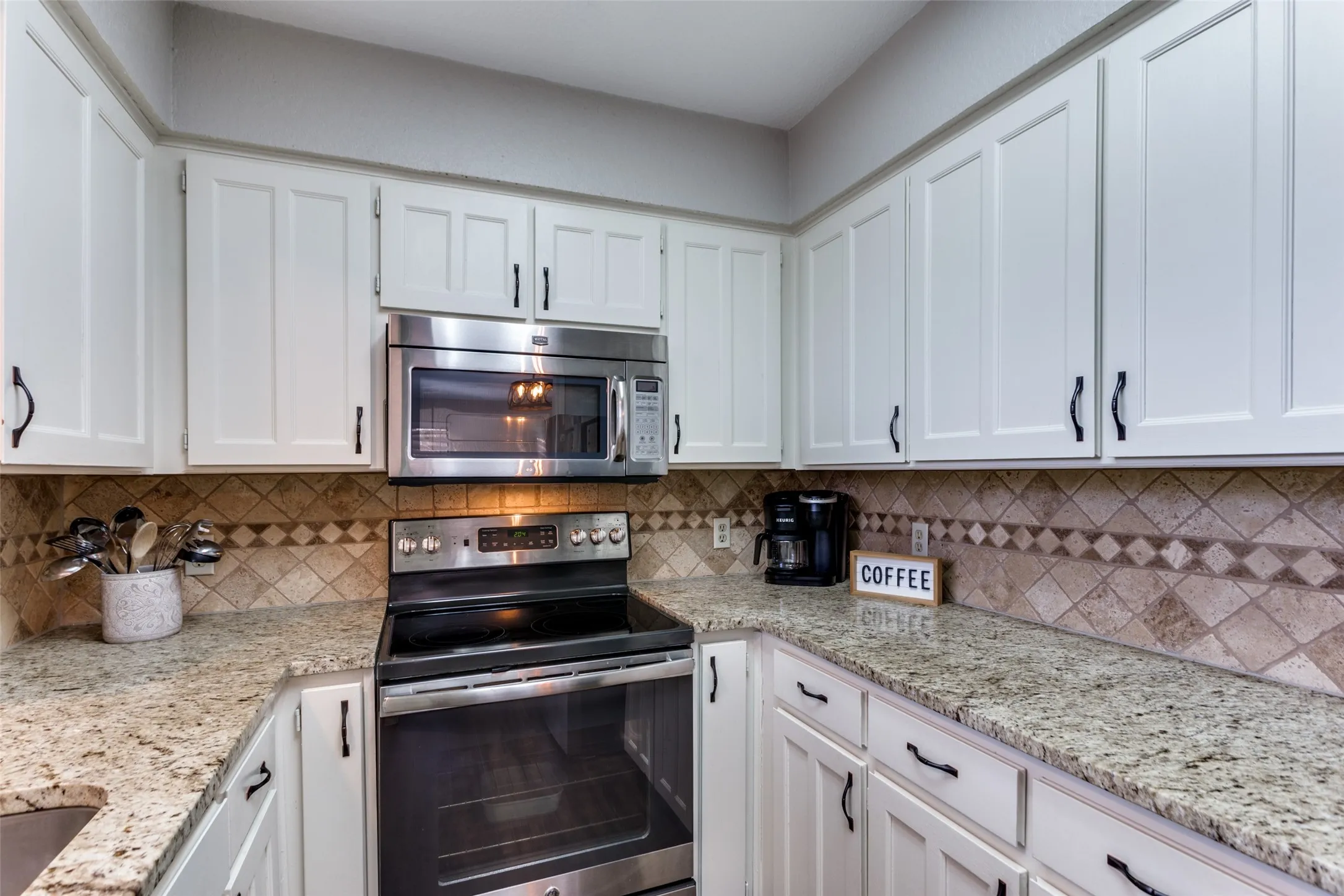 White cabinetry with black hardware, granite countertops, patterned tile backsplash, stainless steel microwave, and a stainless steel range