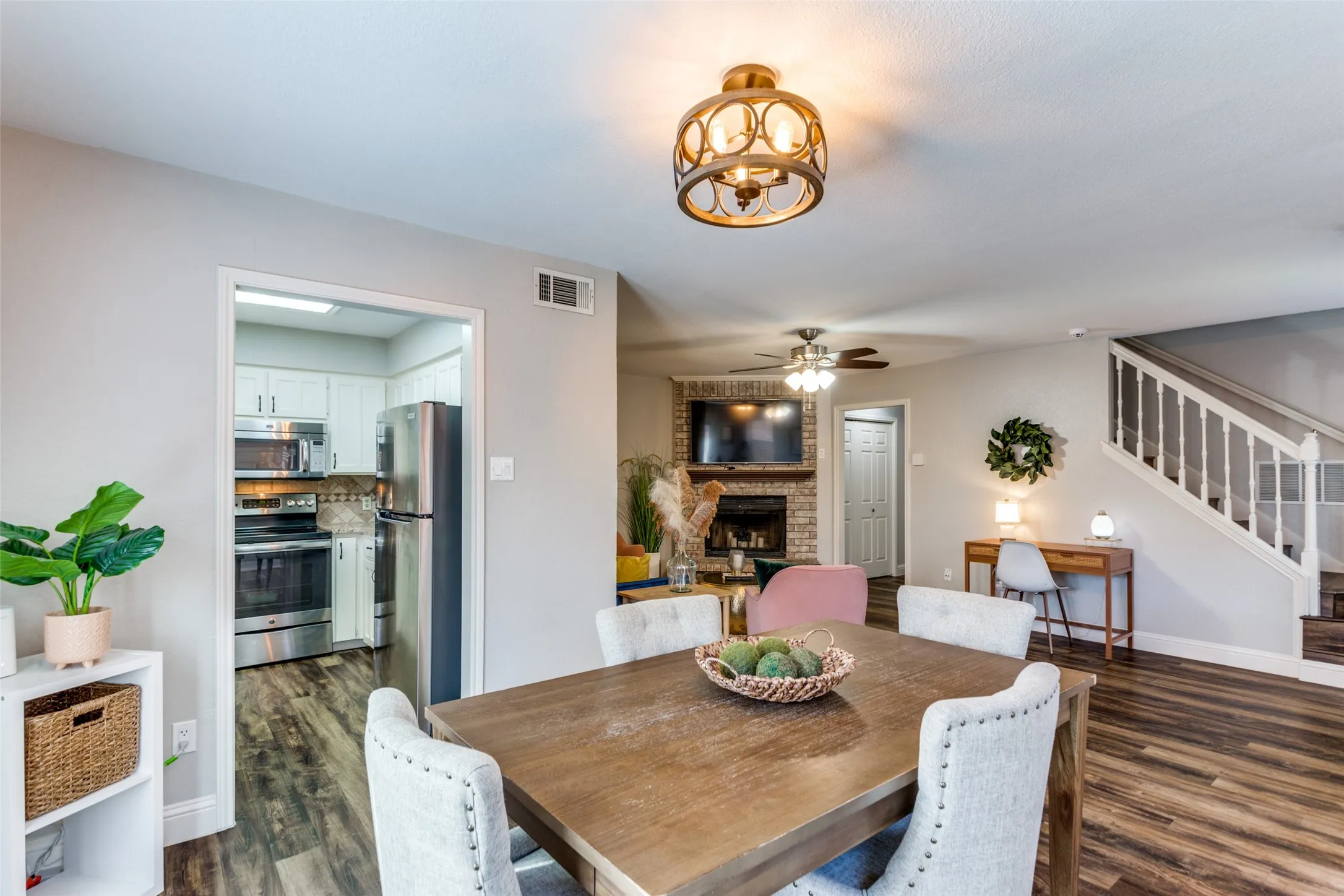 Wood-finish flooring throughout, living area with brick fireplace, staircase with white spindles, and a kitchen featuring stainless steel appliances