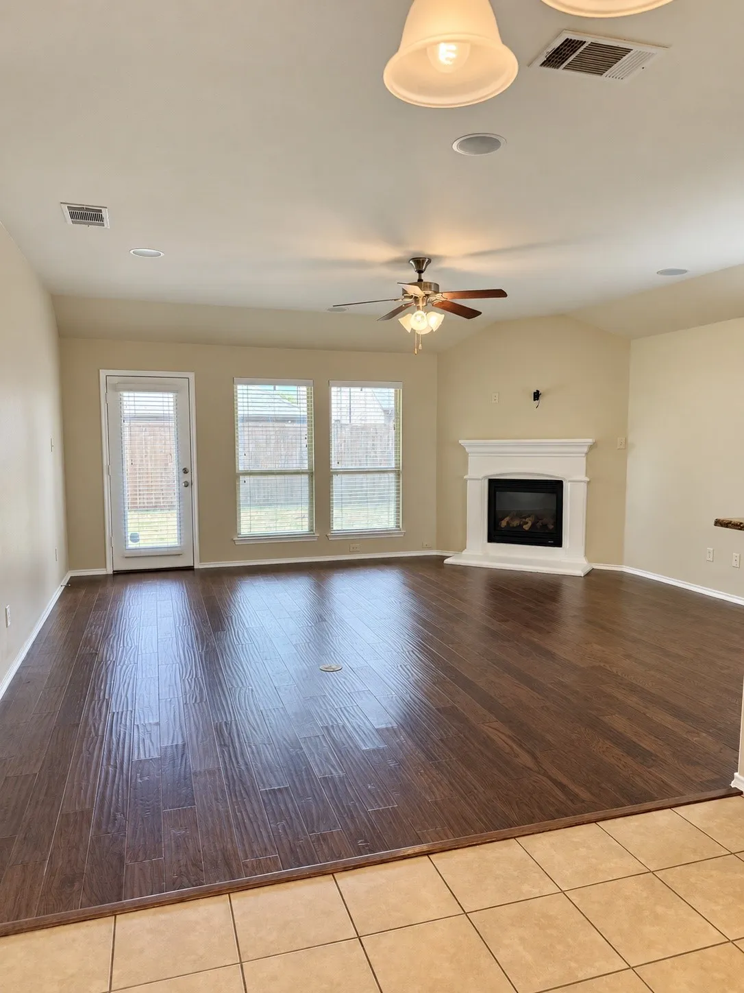 Spacious living area featuring wood-finish flooring, a ceiling fan, three windows, and a fireplace with a white mantel