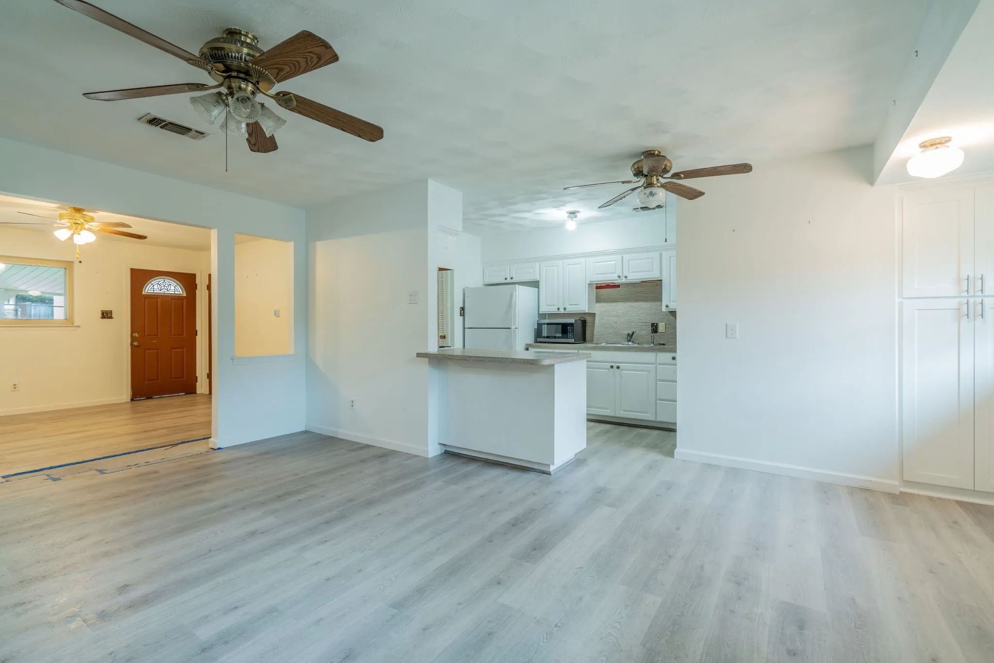 Light wood-finish flooring throughout, white painted walls, ceiling fans, white kitchen cabinetry, and a wood entry door with an arched window