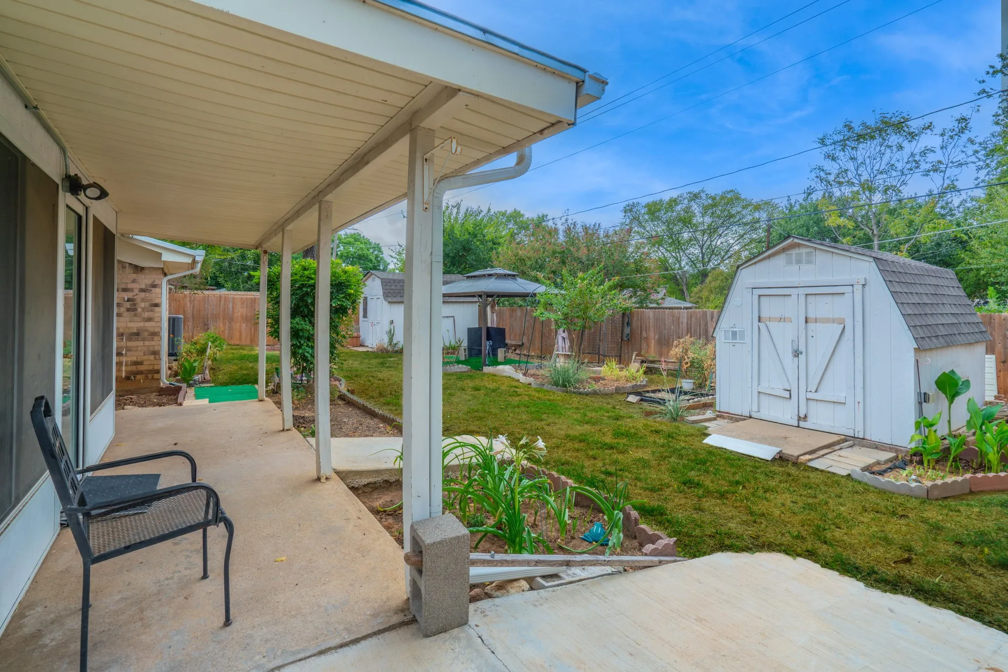 Covered concrete patio, expansive lawn, white storage shed with double doors, wood privacy fencing, and mature landscaping