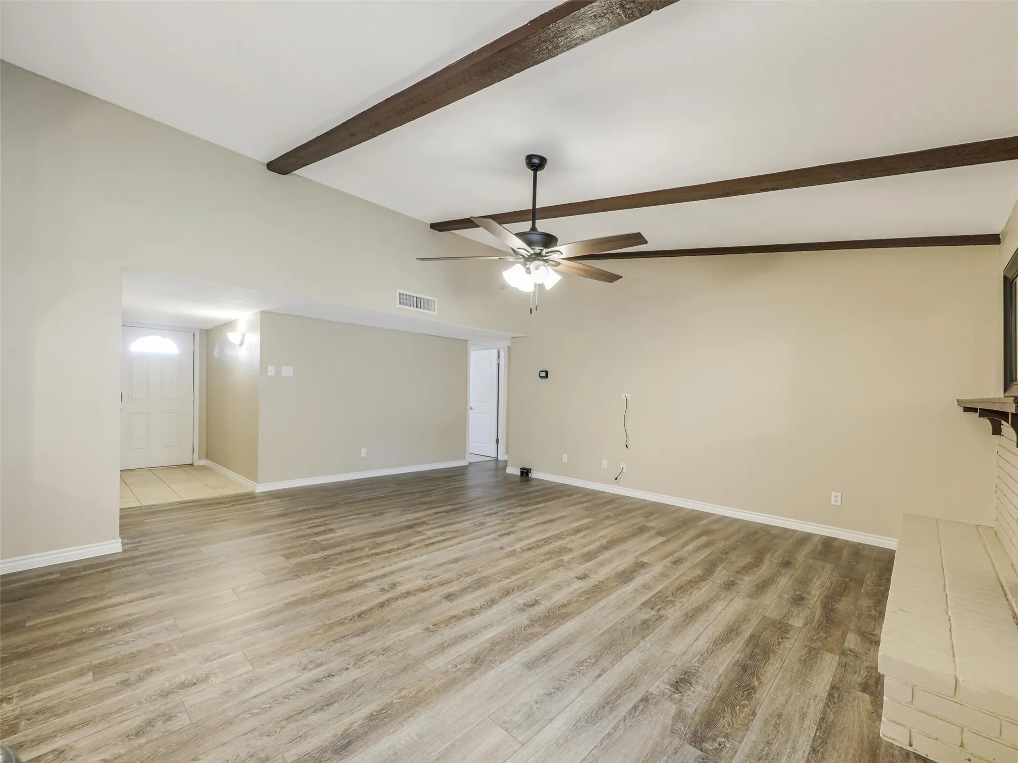 Vaulted ceiling with exposed wood beams, wood-finish flooring, ceiling fan with integrated lighting, brick fireplace with mantel, and recessed entry