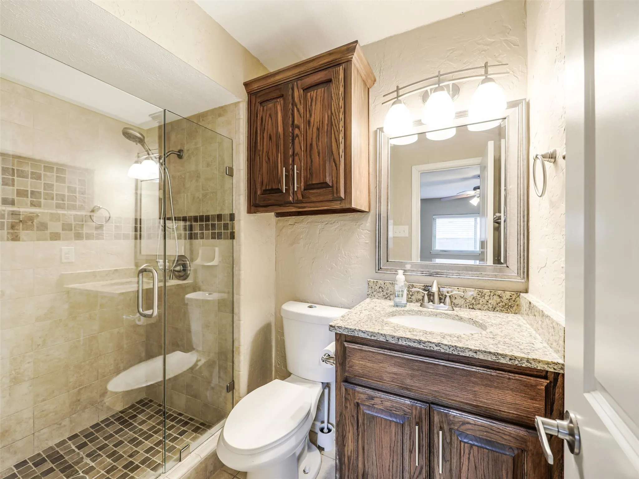 Enclosed shower with full-height tile surround and glass door, wood vanity with a stone countertop and integrated sink, and overhead wood cabinetry