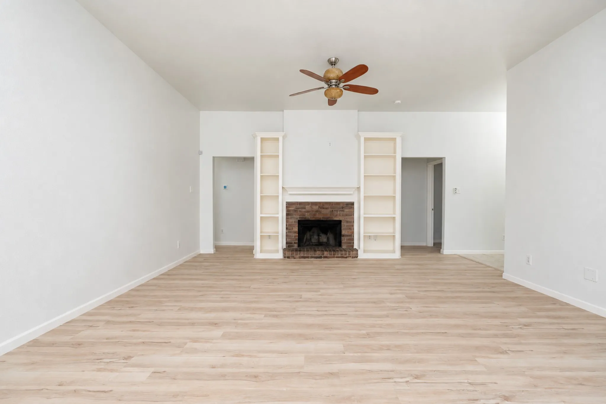 Spacious living area featuring wood-finish flooring, a brick fireplace with a mantel, built-in shelving, and a ceiling fan
