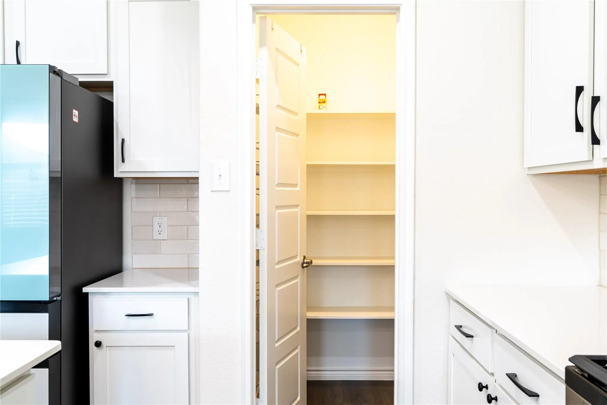 Bright white cabinetry with matte black hardware, white subway tile backsplash, white countertops, and a full-size pantry with shelving