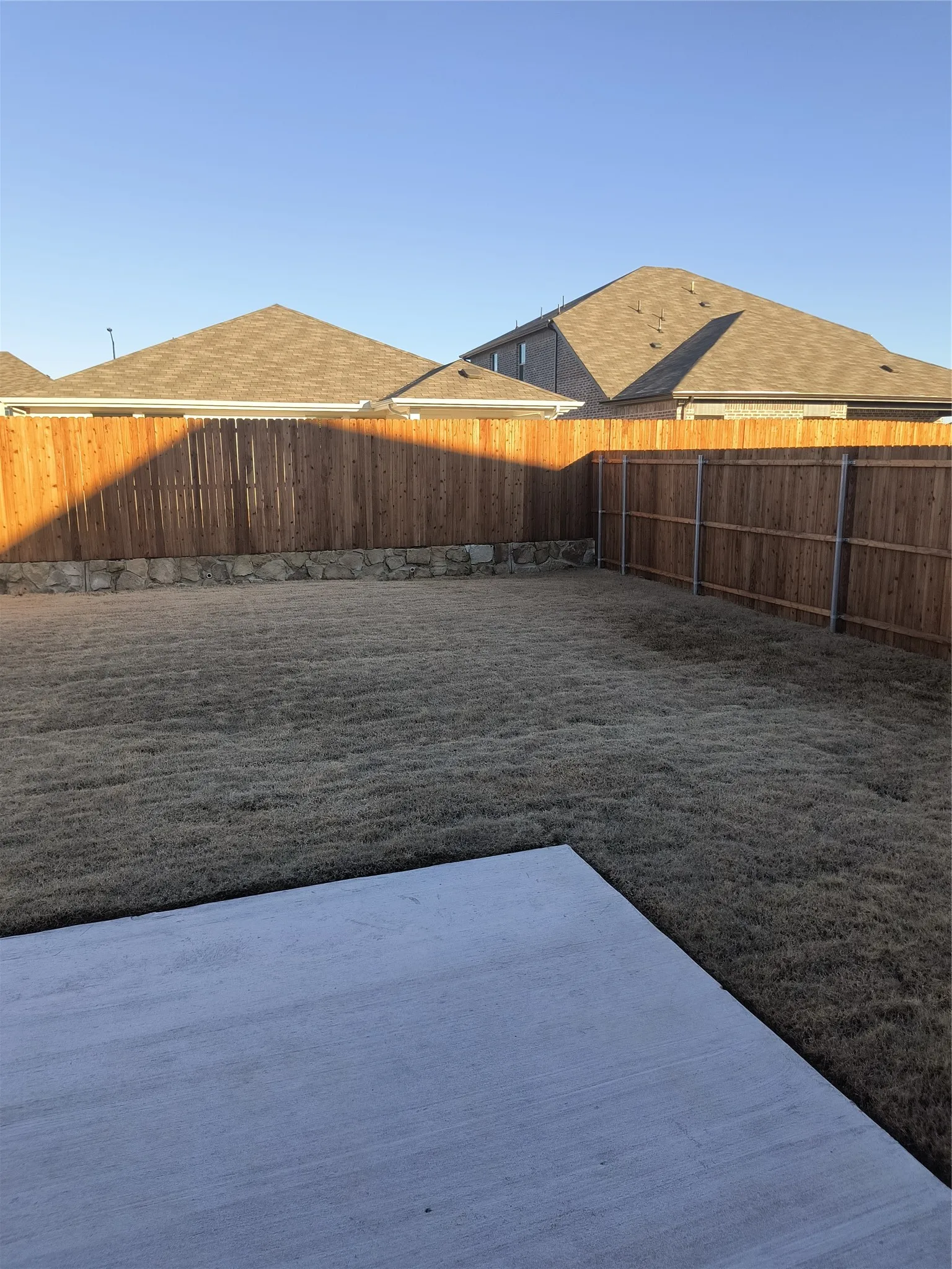 Expansive backyard featuring a concrete patio, wood privacy fencing, a natural stone retaining wall, and a manicured lawn