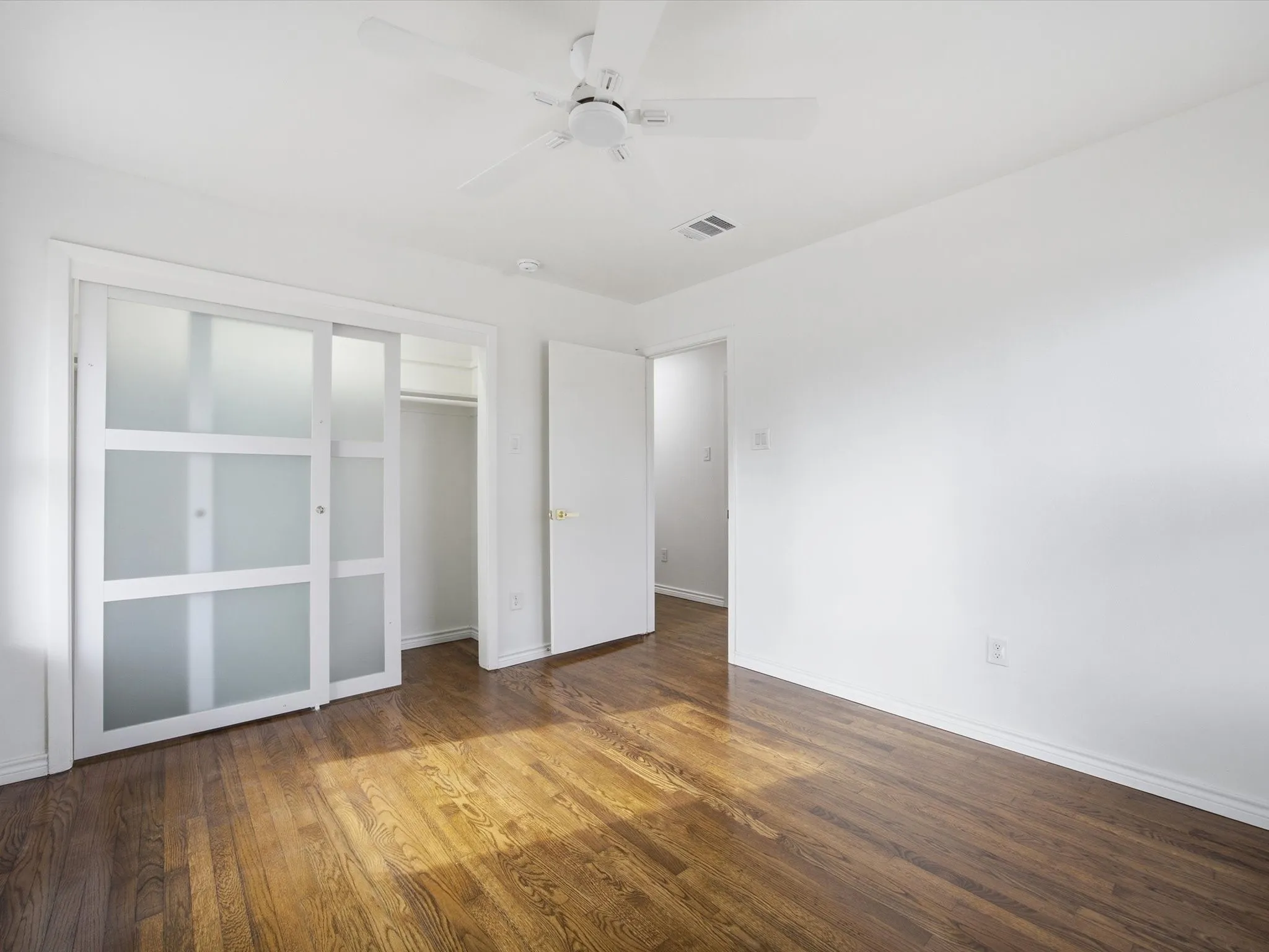 Hardwood flooring throughout, white walls, white ceiling fan, and sliding closet doors with frosted glass panels